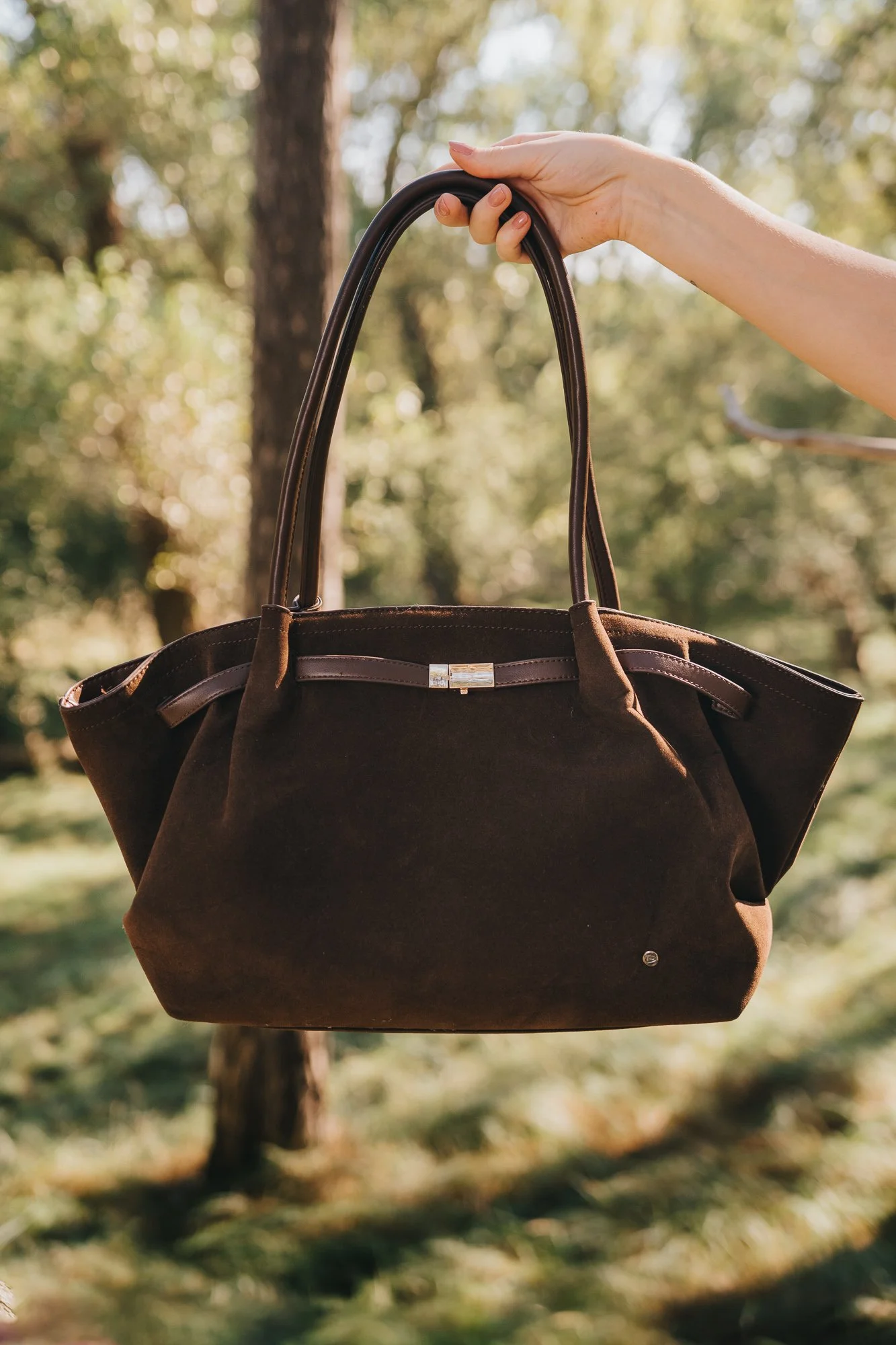 A person holding a brown handbag outdoors among trees, with the sunlight filtering through the leaves.