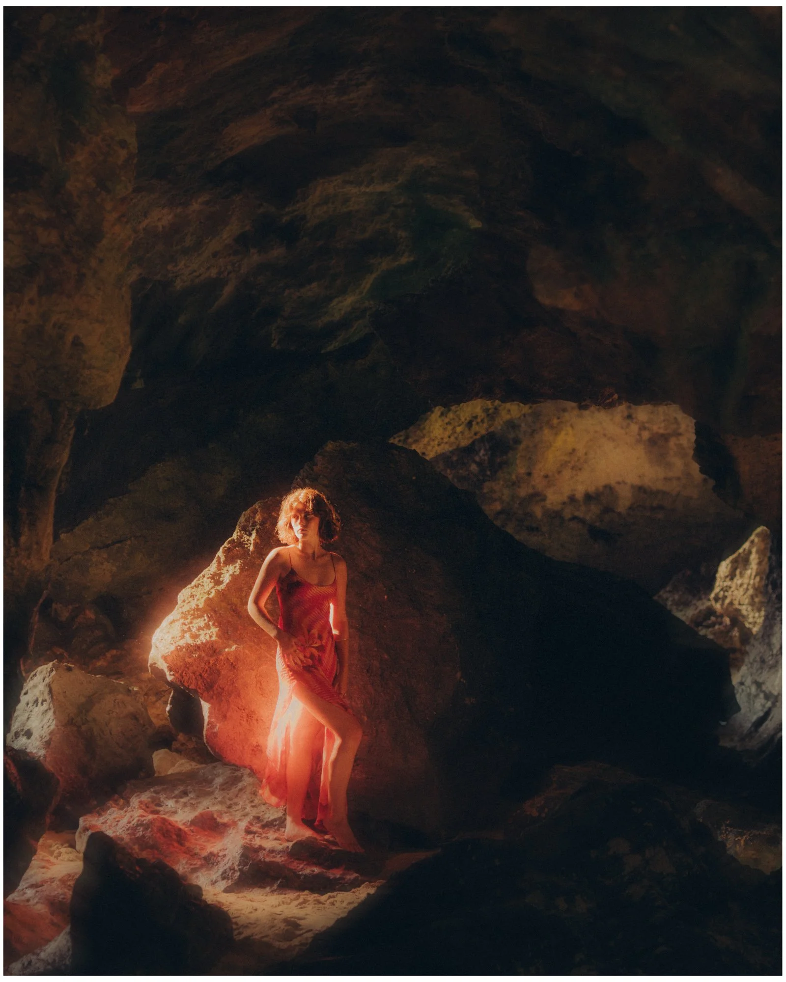A woman in a flowing, red dress standing on rocks inside a dark cave, with light illuminating her and the surrounding rocks.