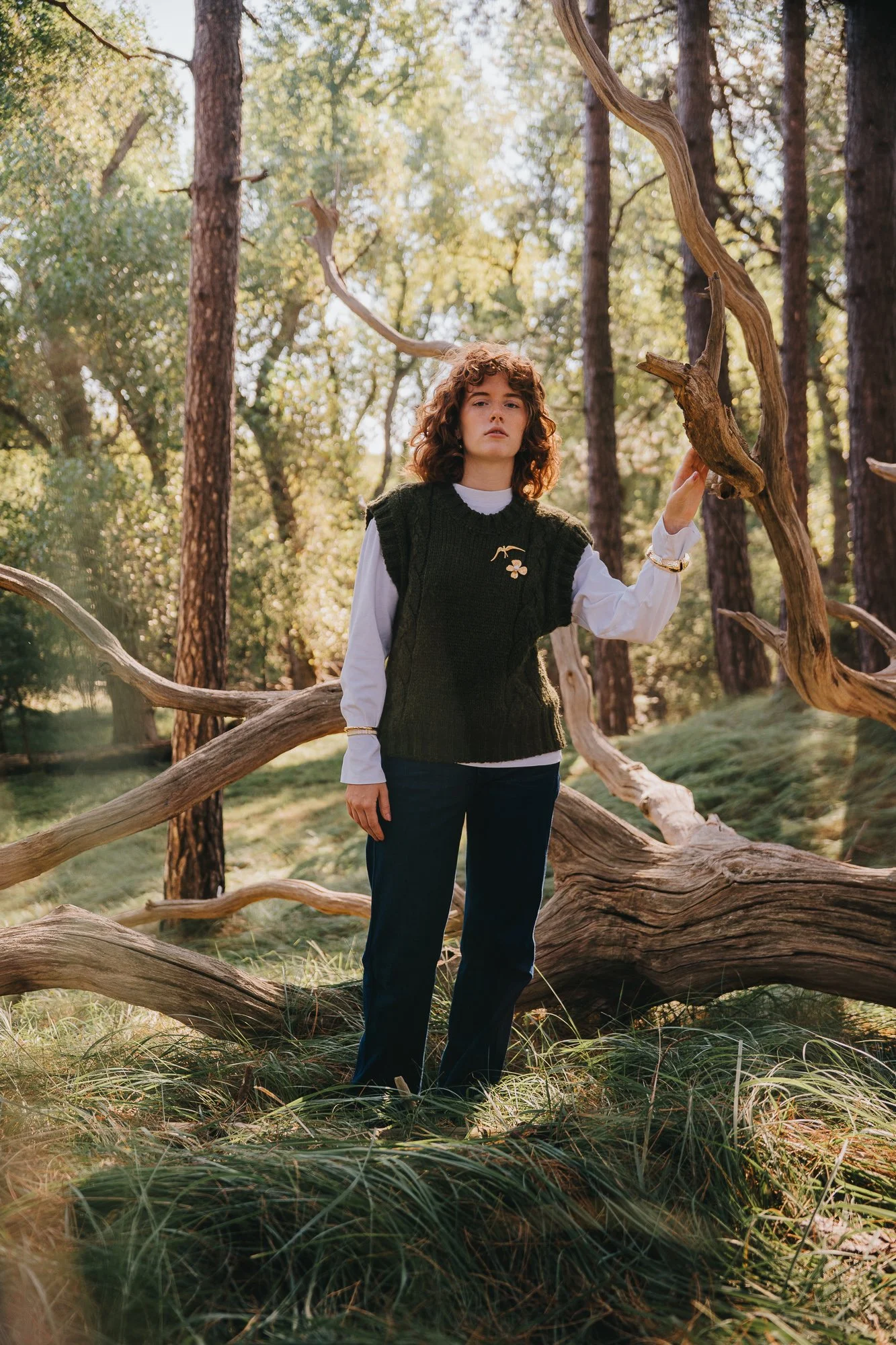 A woman with curly hair wearing a white shirt, dark vest with a mushroom embroidery, and dark pants, stands in a forest with a fallen tree and tall trees around her, holding a twisted branch.