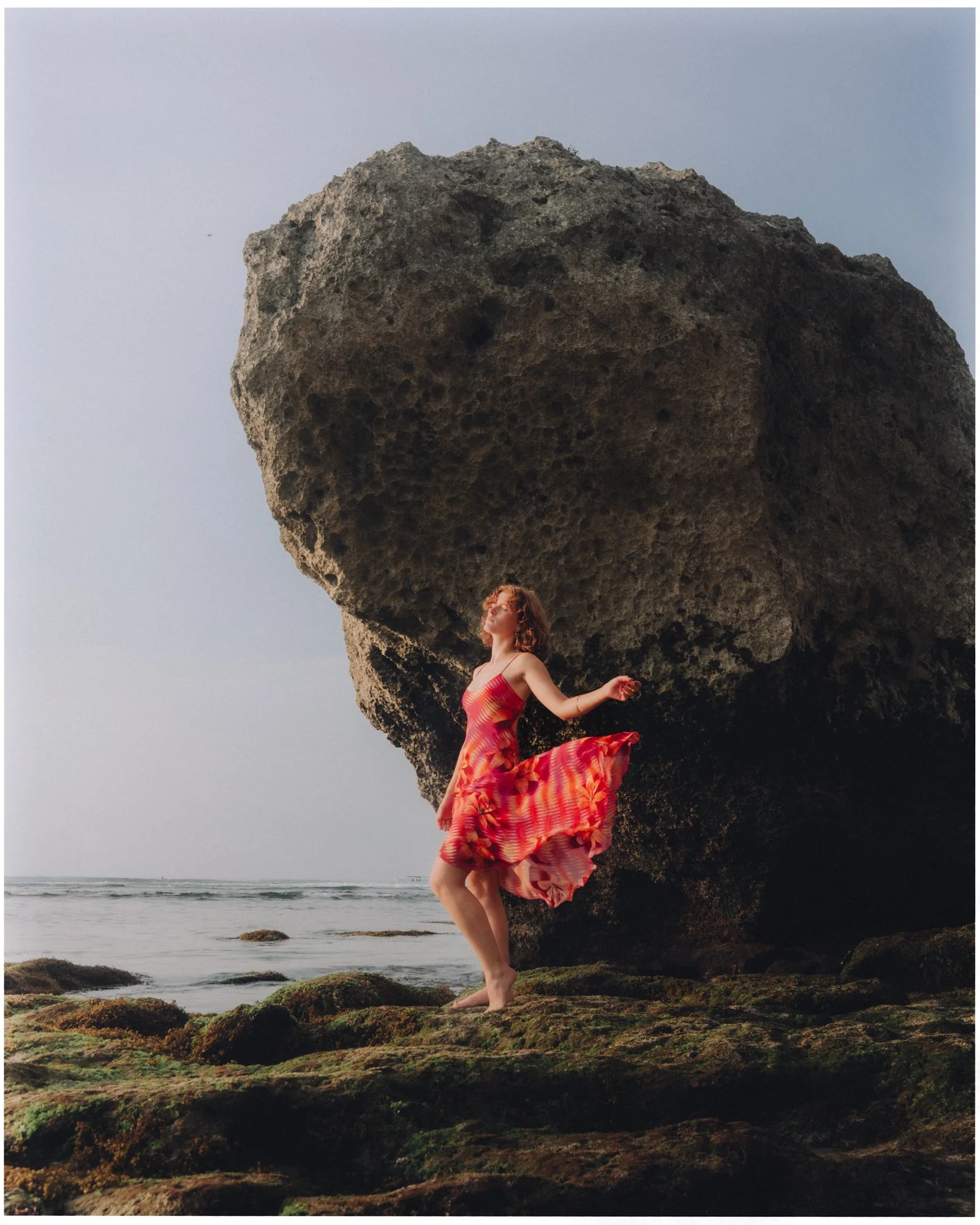 A woman in a colorful red and orange dress standing on mossy rocks at the beach, with a large rock formation and the ocean in the background.