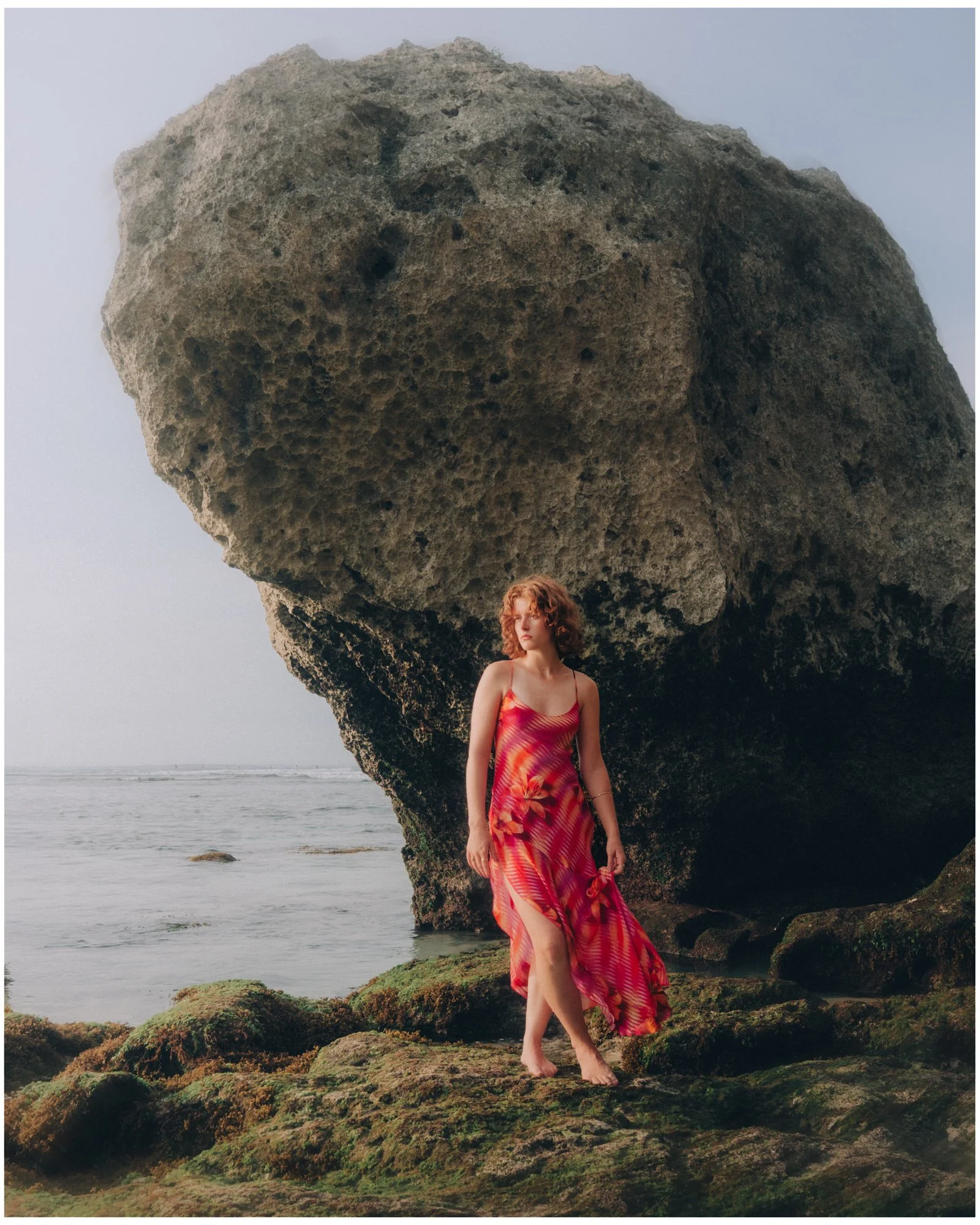 A woman in a colorful pink and orange dress standing barefoot on mossy rocks by the ocean with a large boulder behind her.