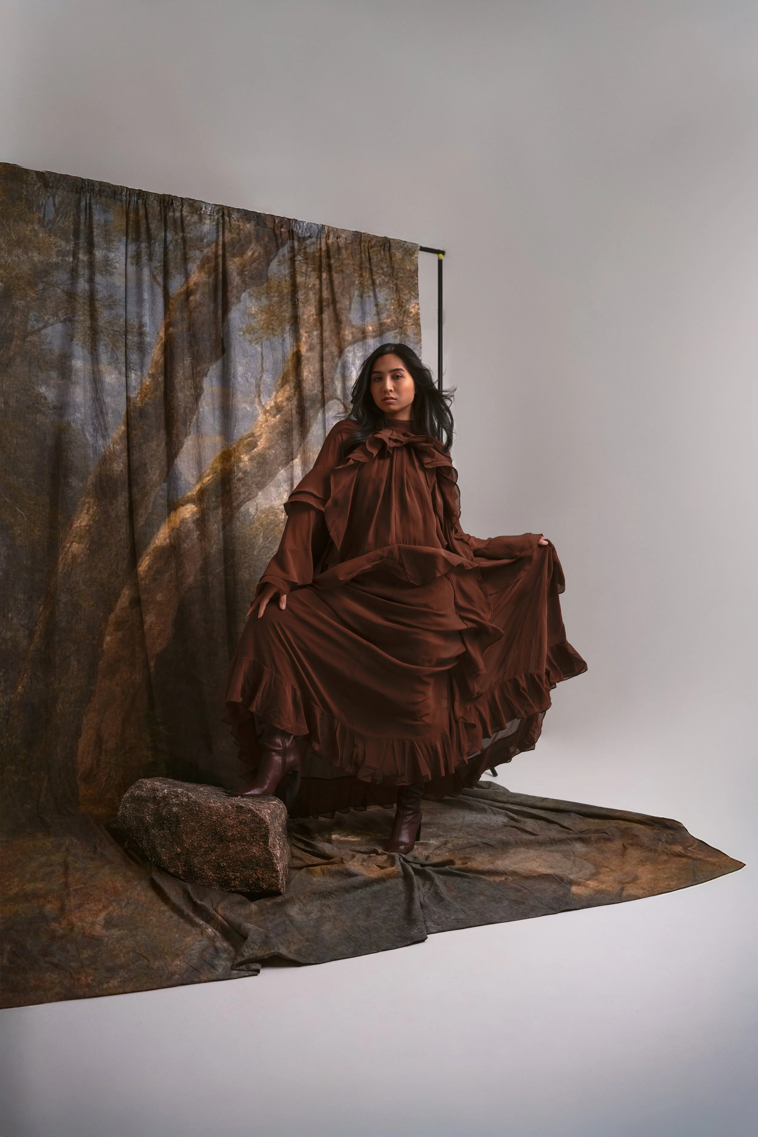 A woman in a long brown dress and matching boots poses on a brown fabric backdrop with a tree scene, standing on a rock.