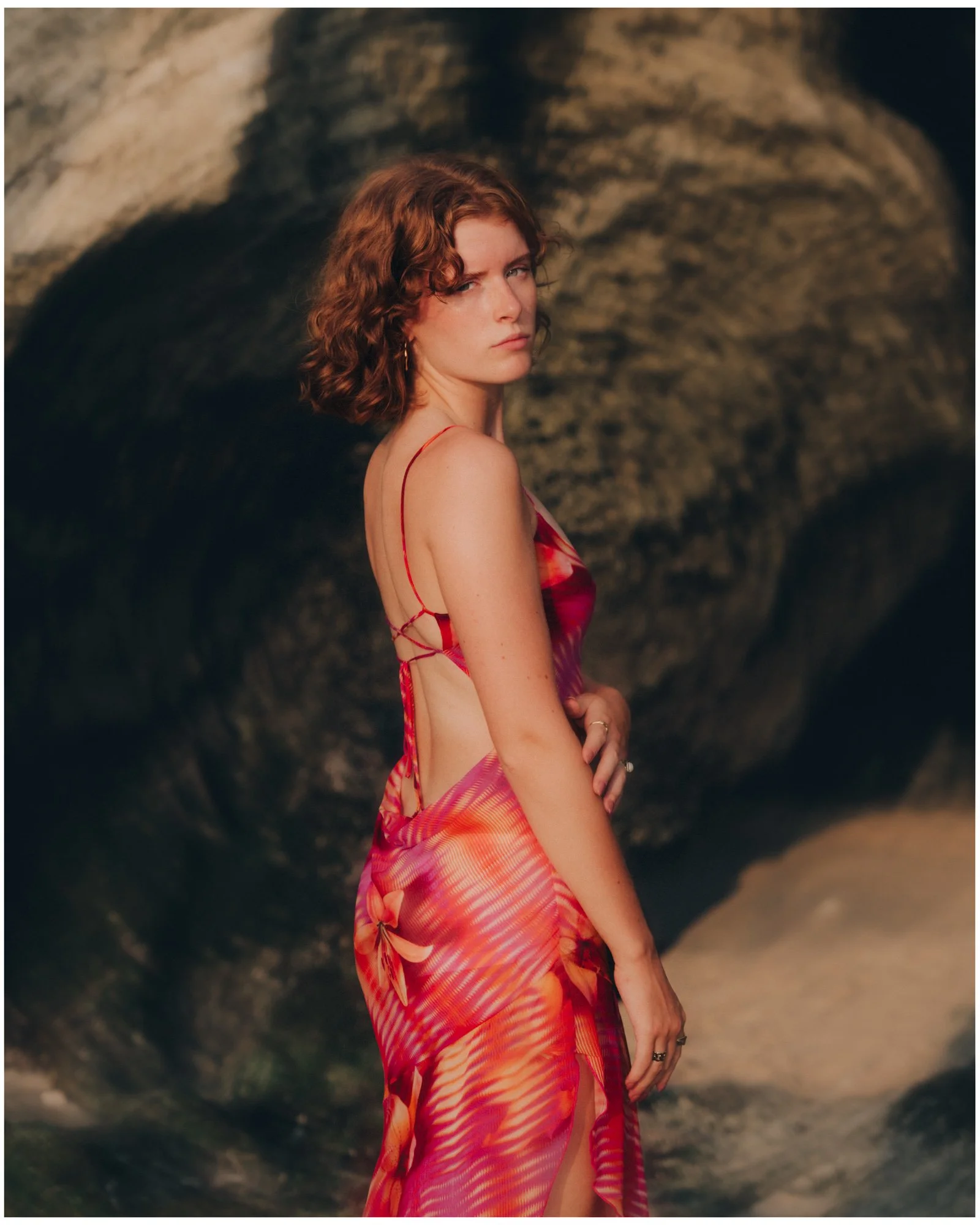 A woman with curly red hair wearing a red and orange colorful dress with open side design, standing in front of a rocky background.