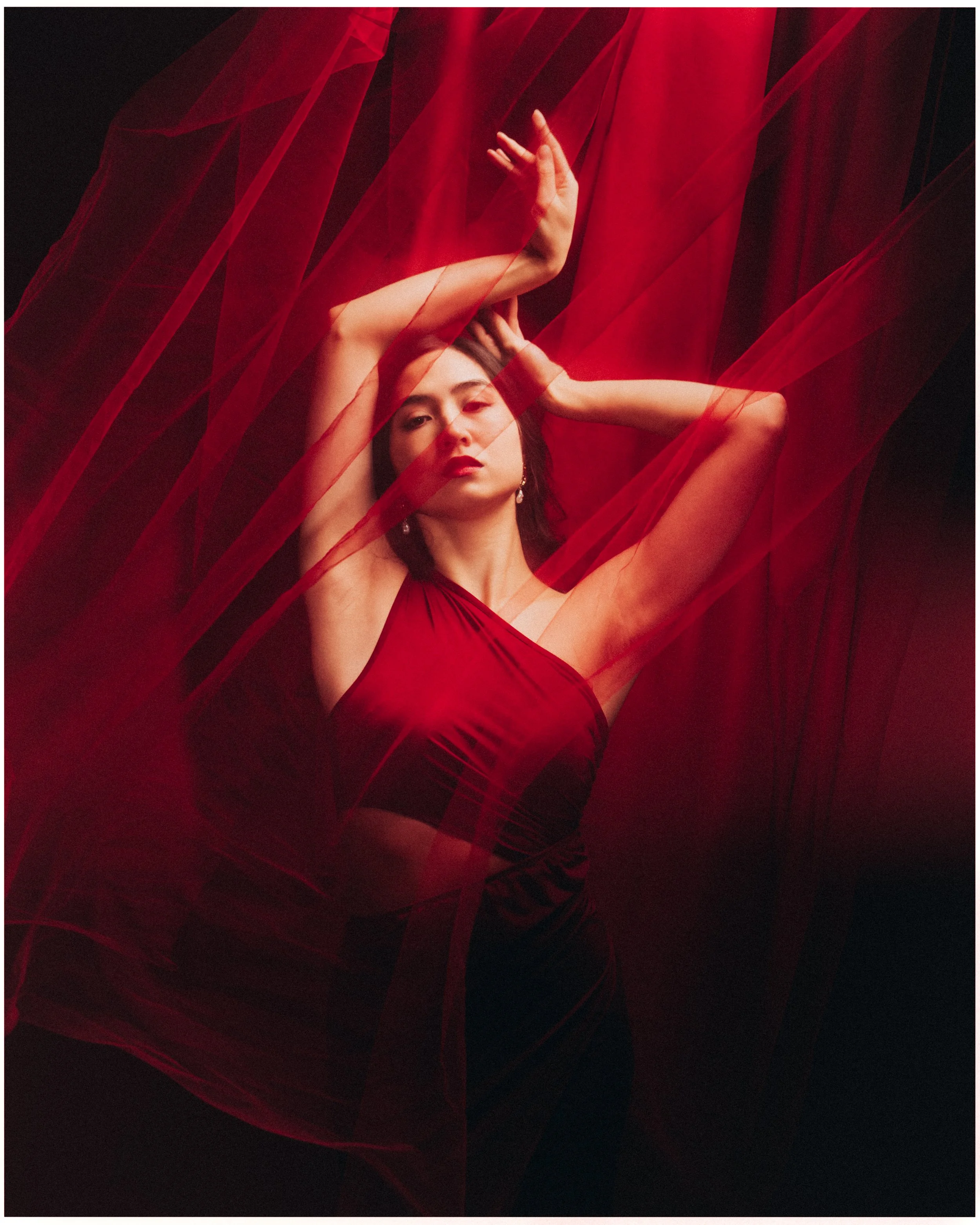 A woman in a red dress poses with her arms raised and intertwined above her head, with sheer red fabric swirling around her against a dark background.