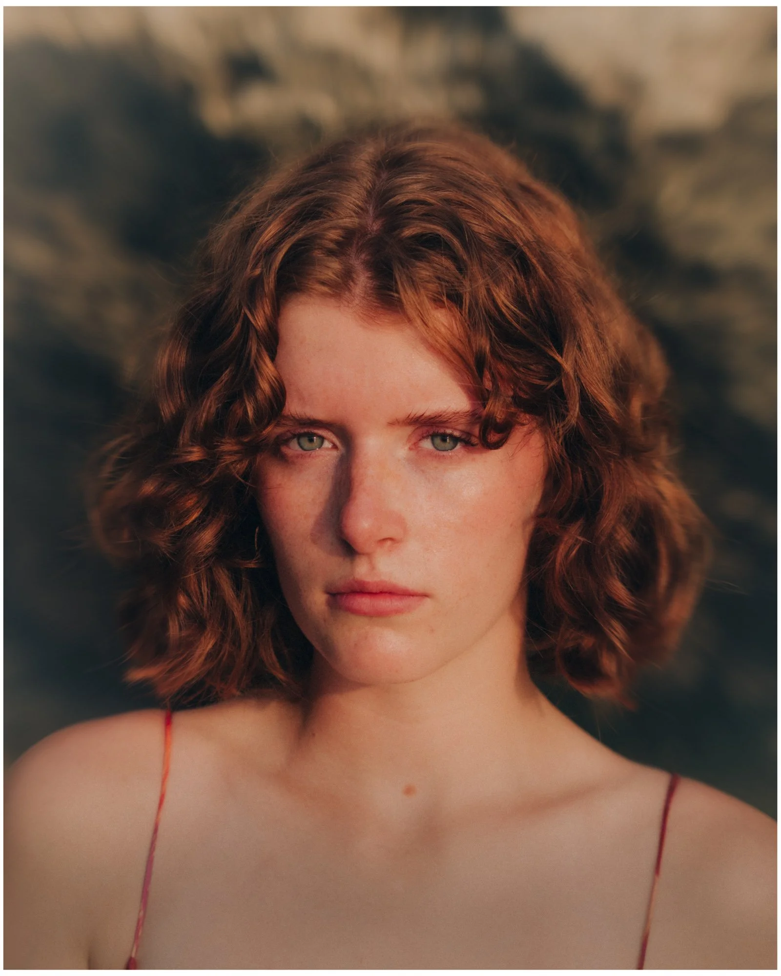 A close-up portrait of a young woman with curly red hair and green eyes, wearing a red spaghetti strap top, with a blurred stone background.