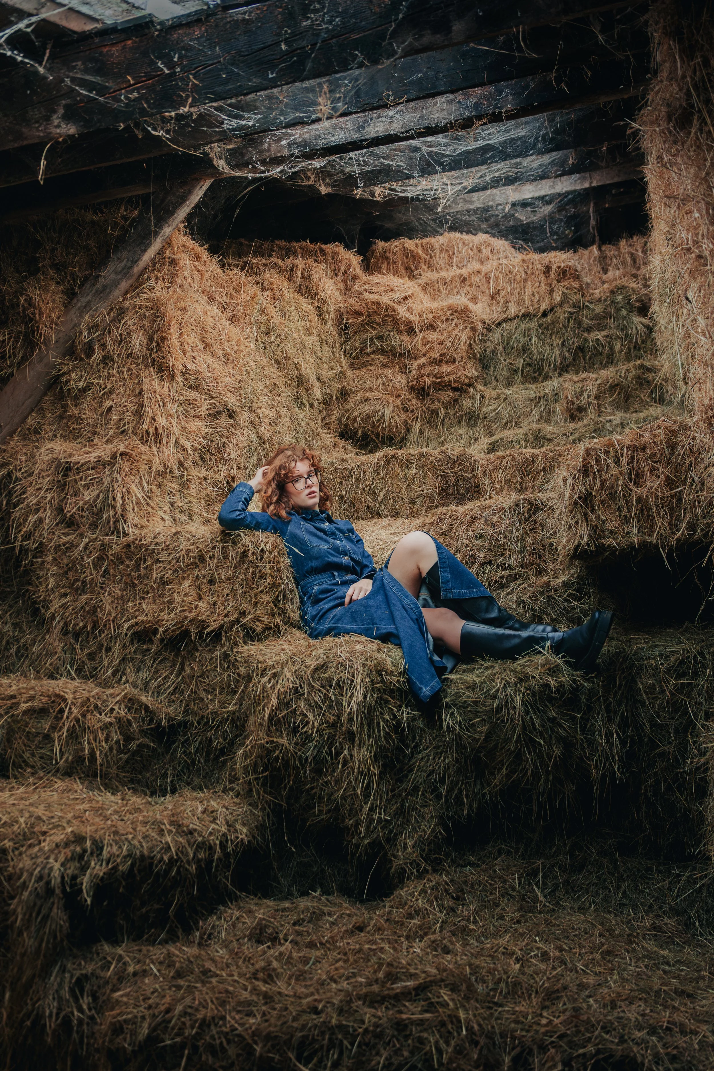A woman in a blue denim dress and black boots relaxing on a pile of hay bales inside a barn.