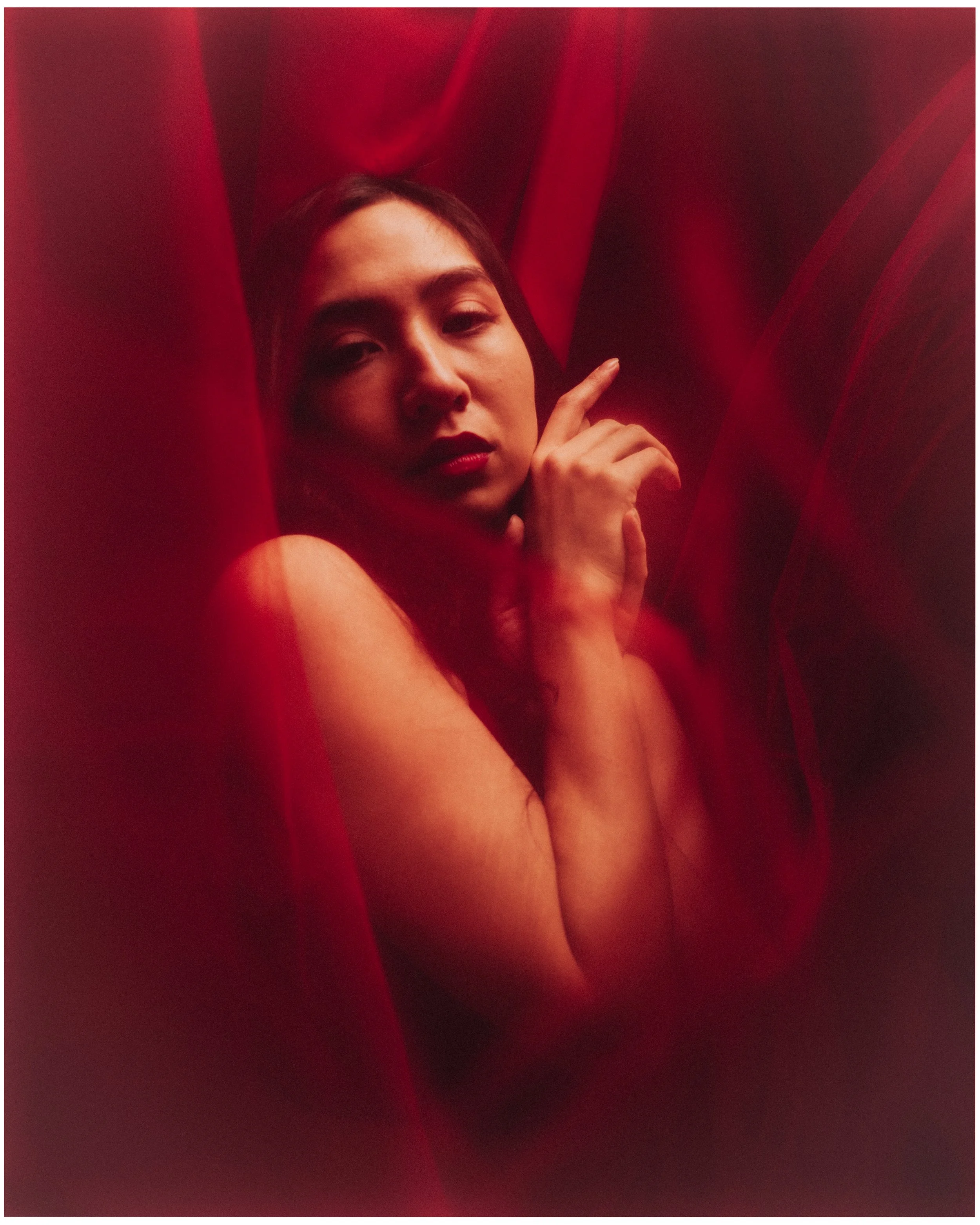 A woman with dark hair and red lipstick is surrounded by red fabric, creating a soft, mysterious atmosphere.