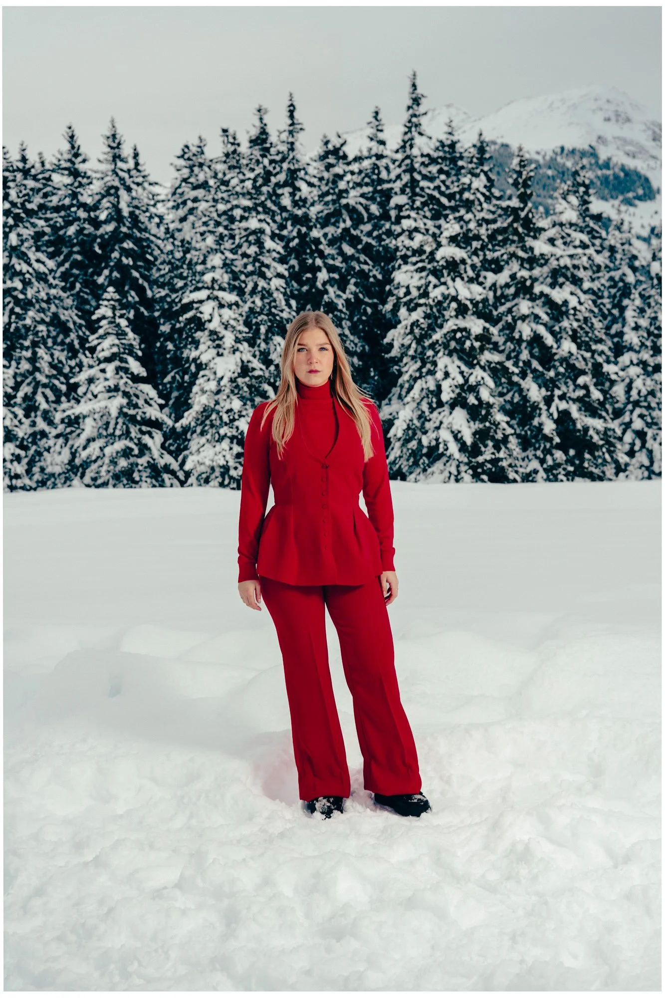 A woman dressed in a red outfit standing in snow with snowy pine trees and mountains in the background.