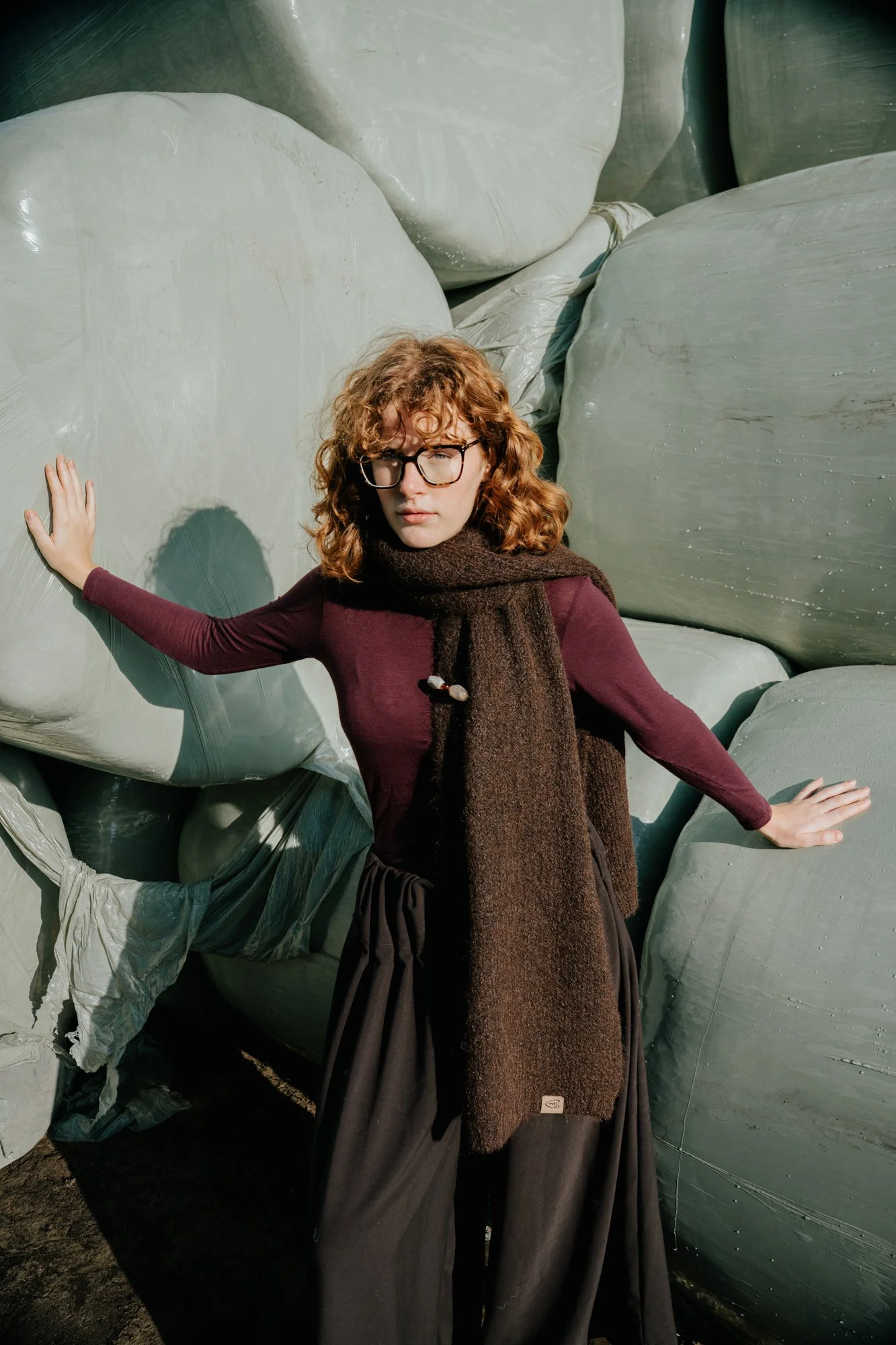 A woman with curly red hair, wearing glasses, a maroon long-sleeved top, a brown scarf, and dark pants, stands between large, green, cylindrical bales of hay or straw, with her hands on the bales, looking at the camera.