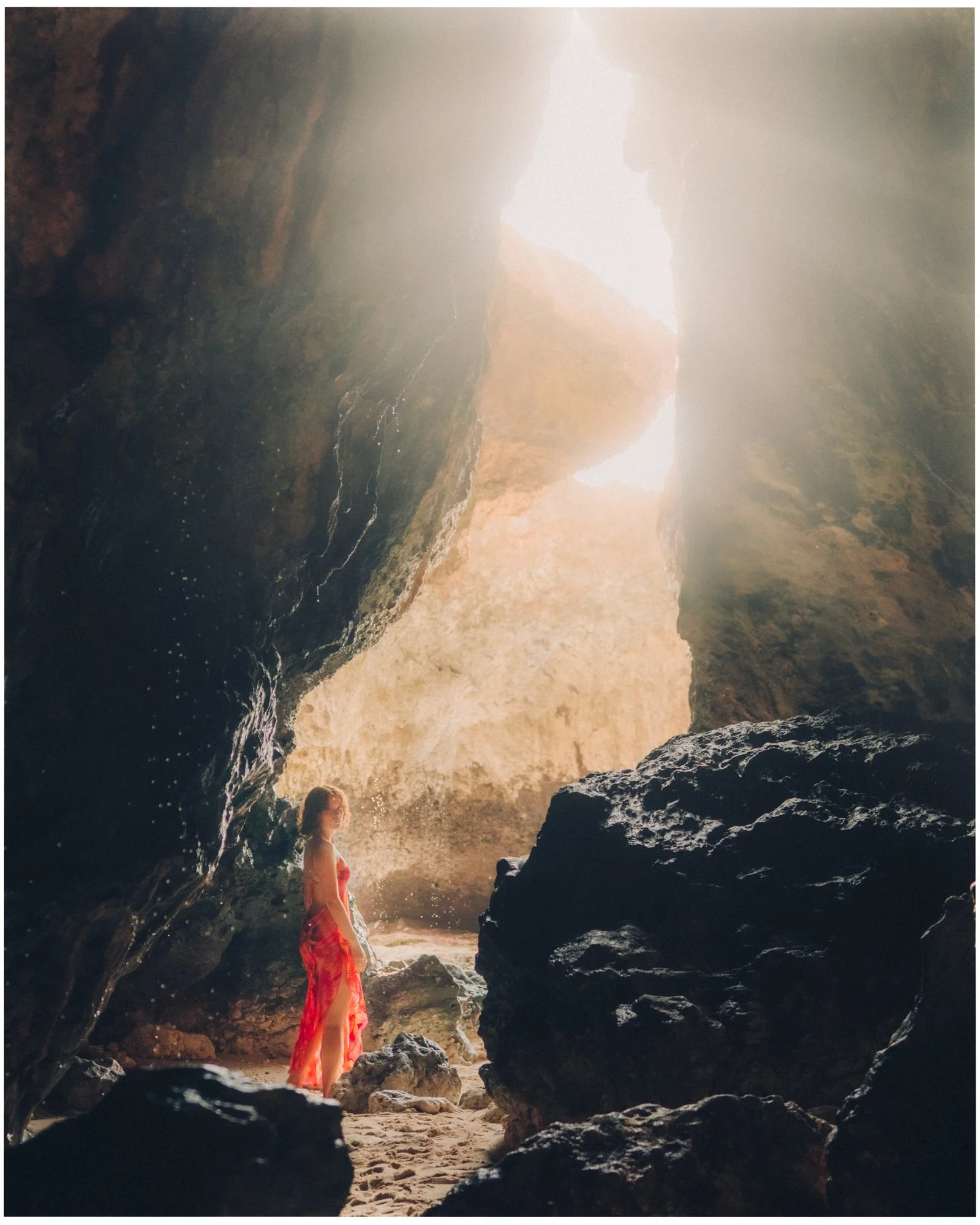 A woman in a red dress standing inside a large, rocky cave with sunlight shining down from above, sandy ground, and water splashing against the rocks.