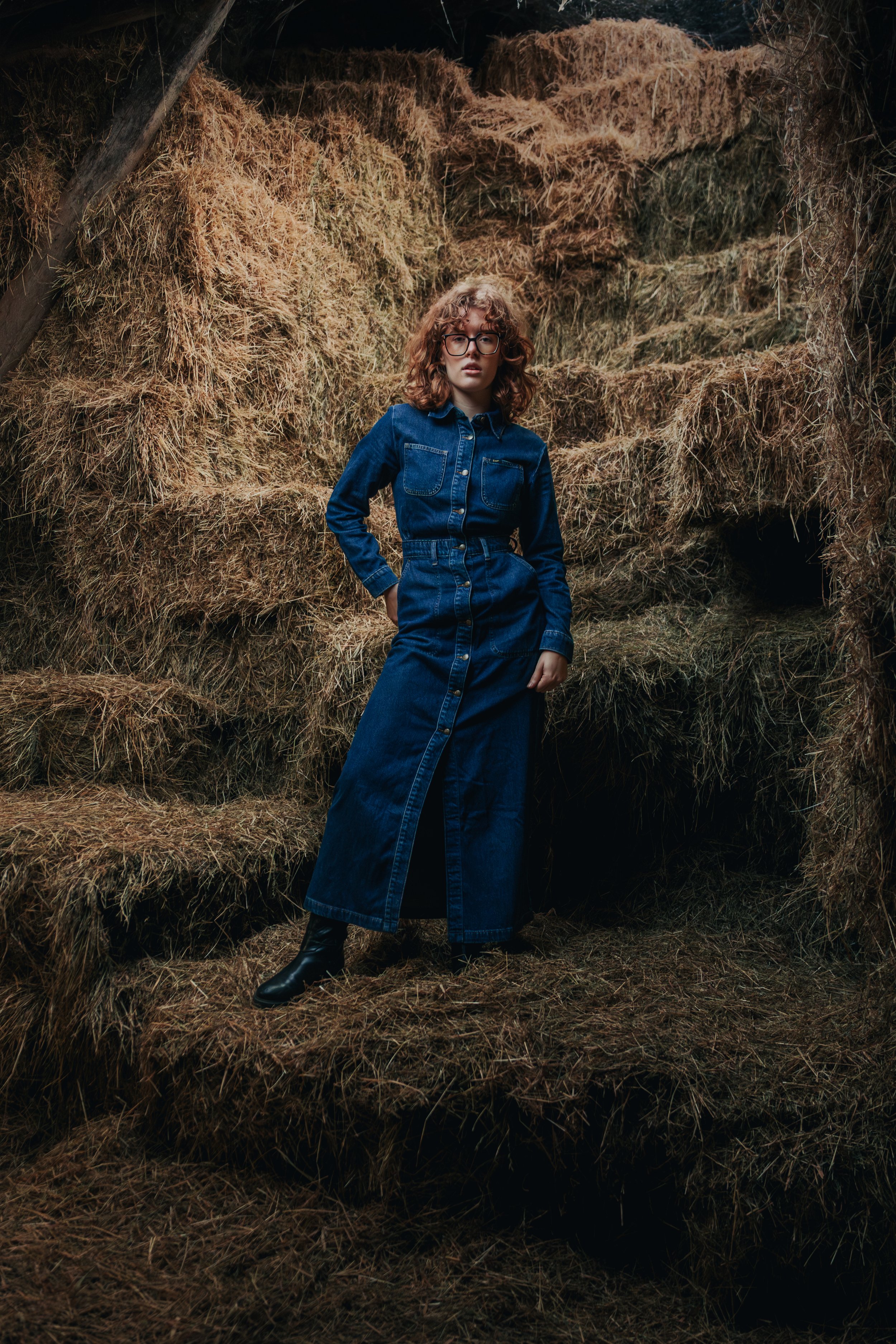 A woman with curly hair and glasses wearing a denim dress and black boots standing in a haystack setting.
