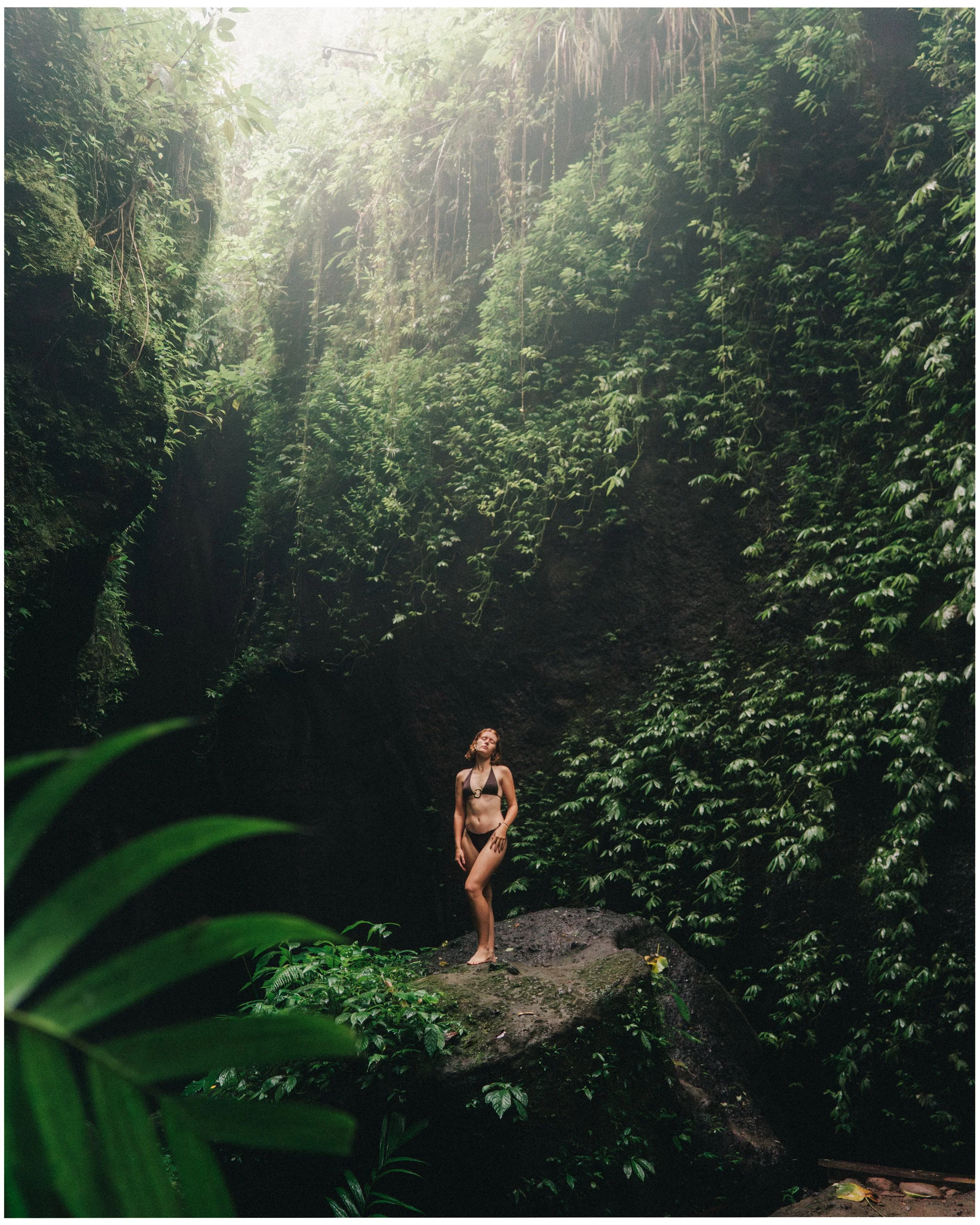 A woman in a bikini standing on a rock in a lush green jungle or rainforest with dense foliage surrounding her.