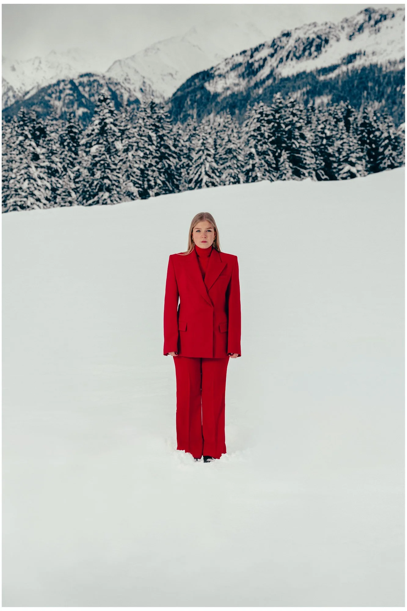 A woman in a red  fashion suit standing in snow with snow-covered trees and mountains in the background.