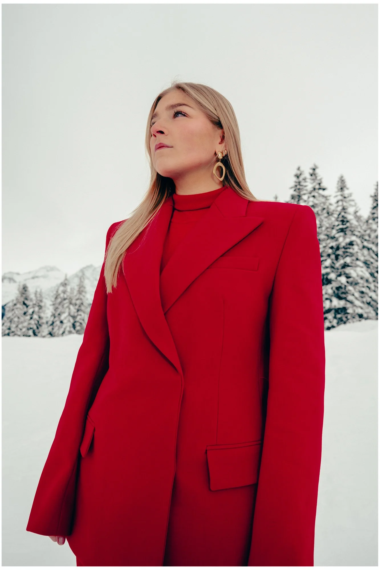 A woman with blonde hair wearing a red suit and gold earrings stands outdoors in a snowy landscape with trees and mountains in the background.