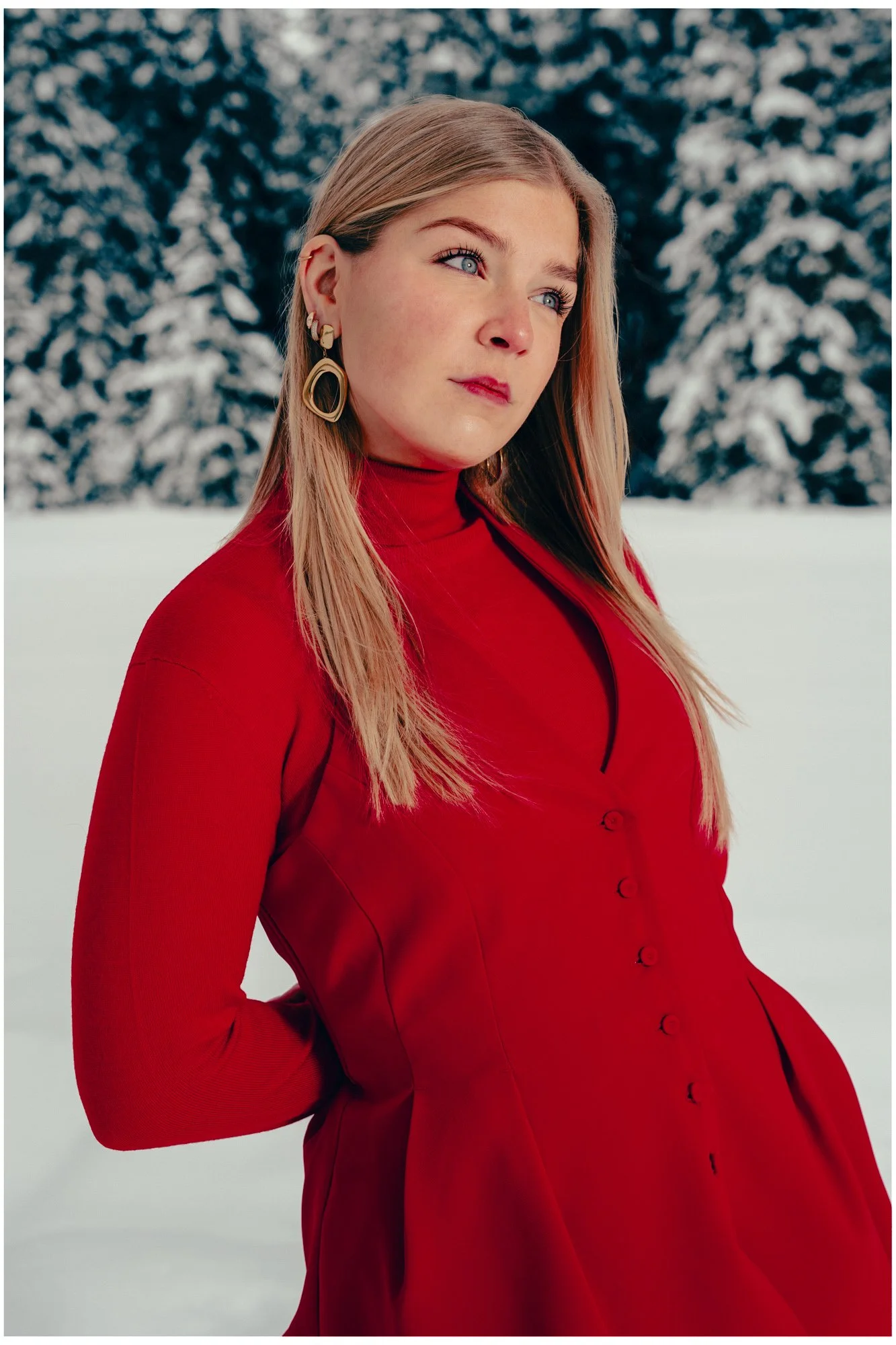 A woman with blonde hair and blue eyes wearing a red dress with a high collar, standing outdoors in a snowy landscape with snow-covered trees in the background.