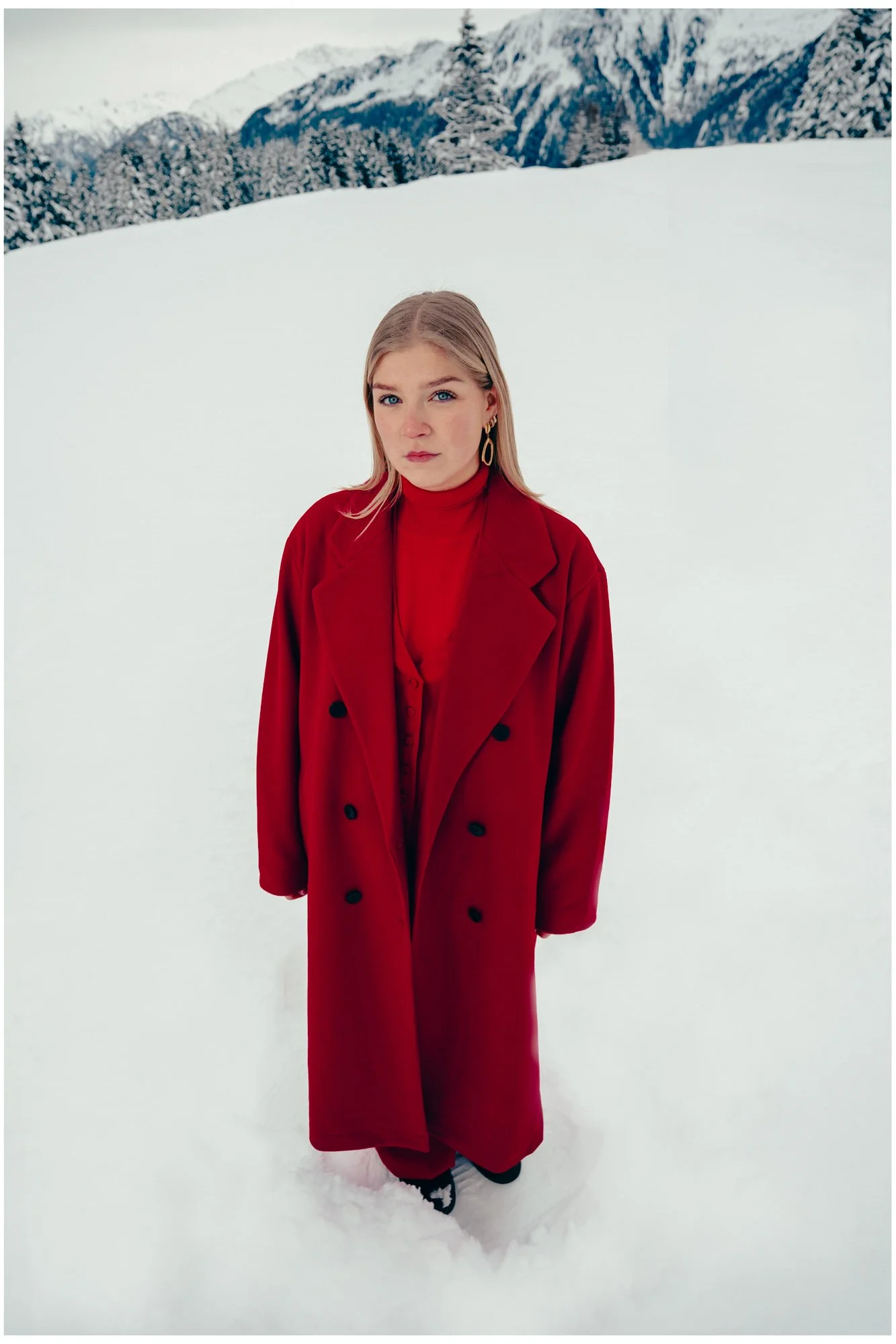 A young woman with blonde hair and blue eyes standing in snow, wearing a red coat and red turtleneck, with mountains and trees in the background.