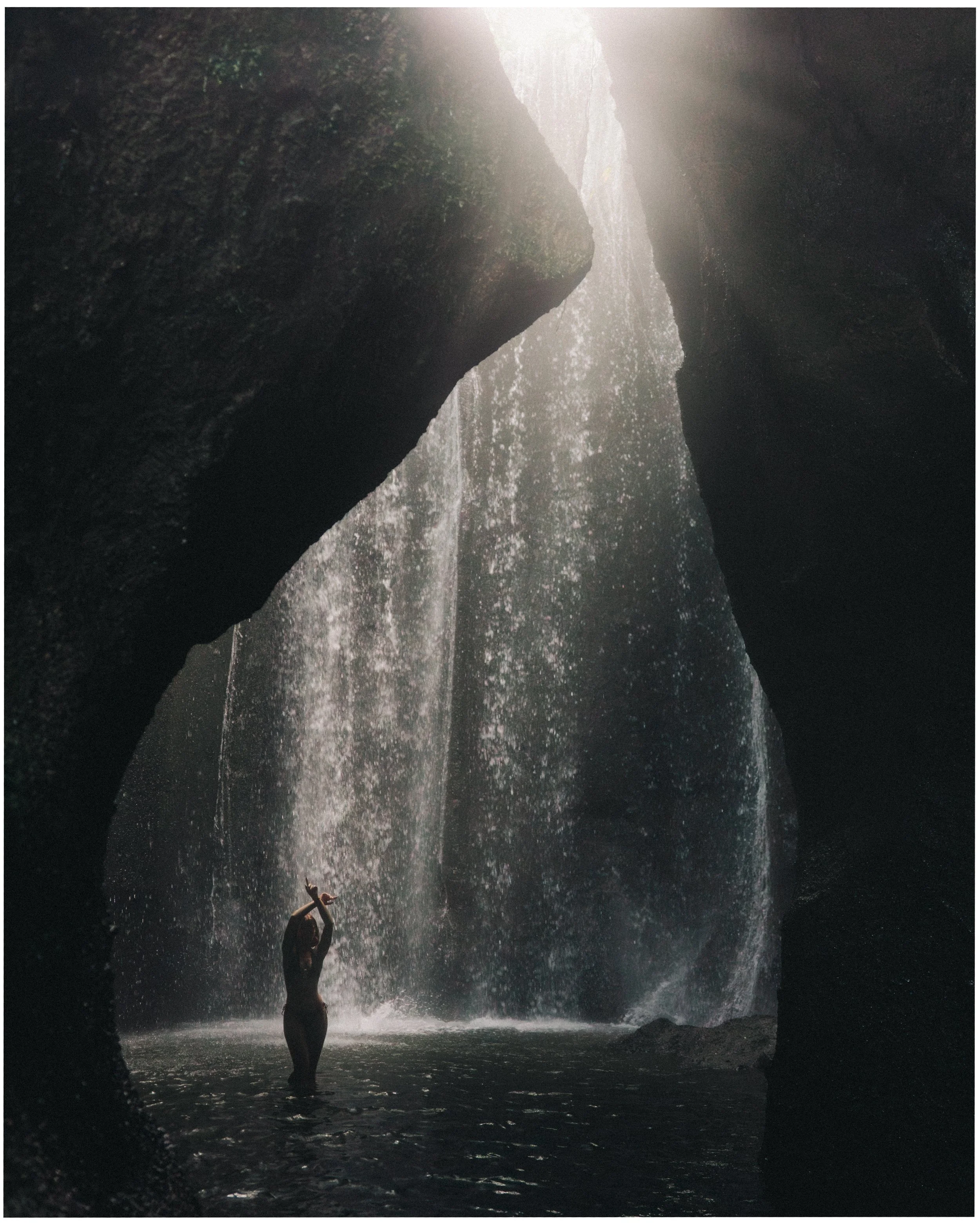 A person standing in a water pool underneath a waterfall, surrounded by dark rock walls and illuminated by sunlight from above.