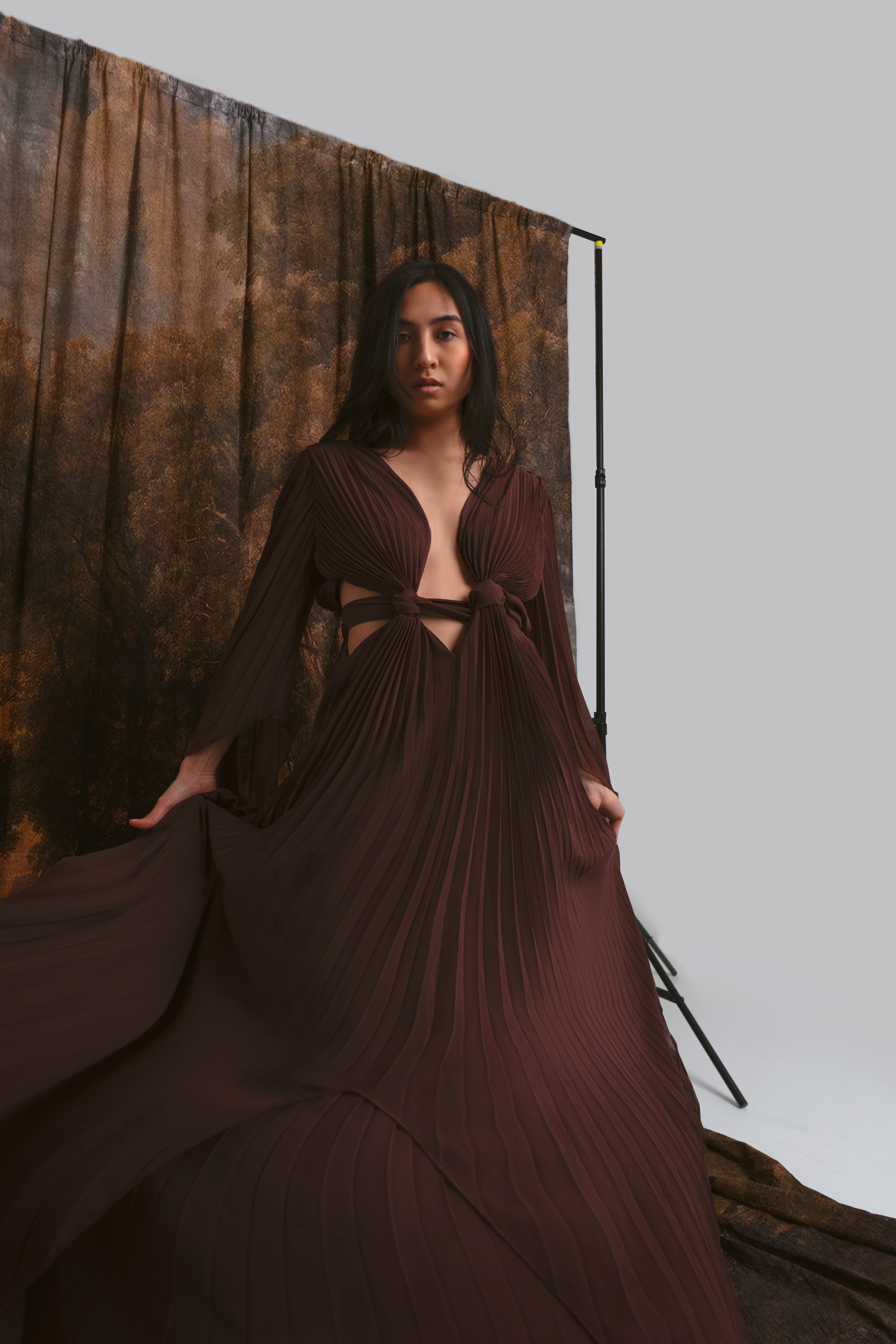 A woman in a flowing, pleated, dark red gown with cutout sides posing in front of a brown textured backdrop on a photography set.