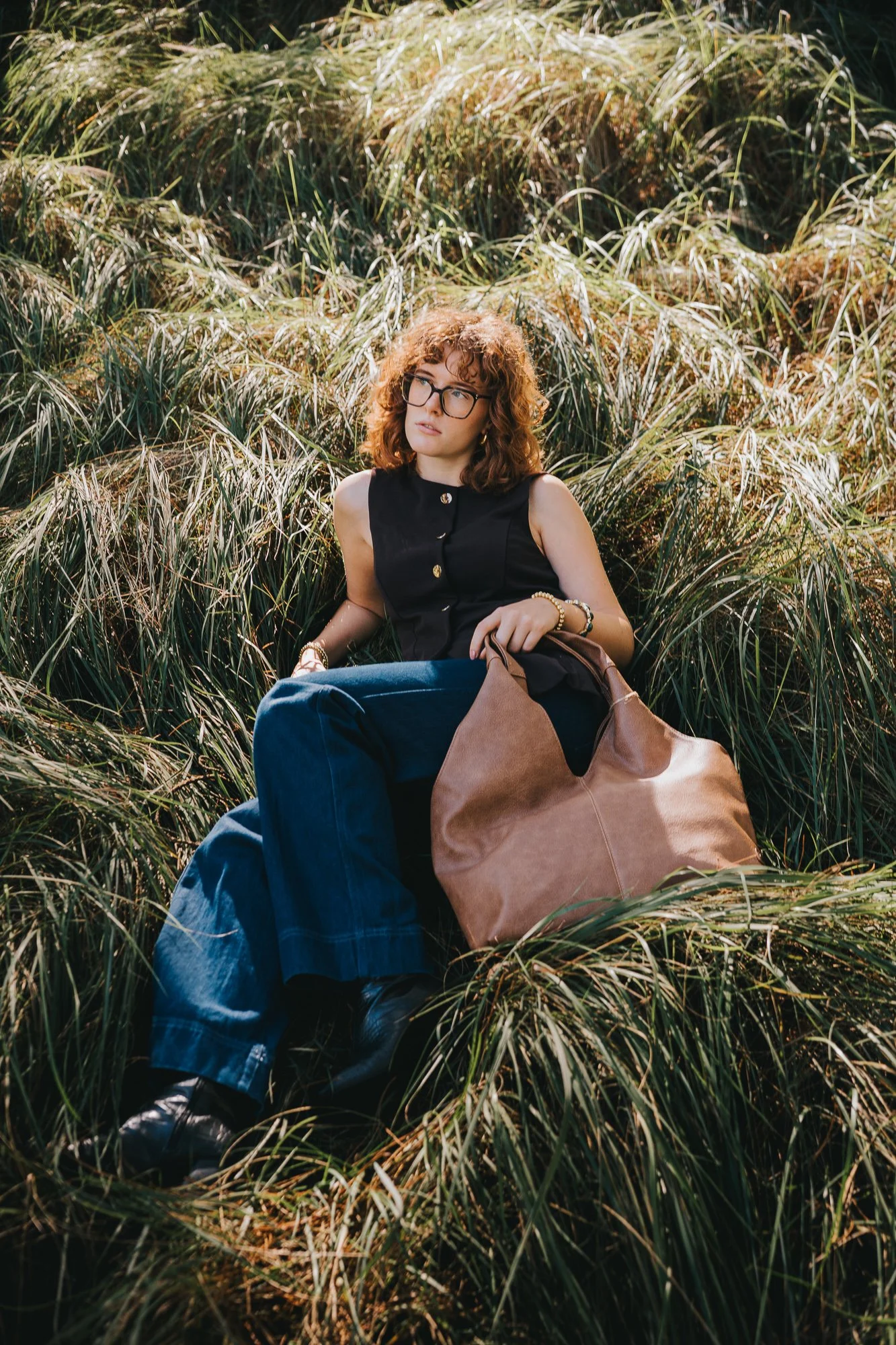 A woman with curly red hair and glasses sitting in tall grass, carrying a large brown leather bag, wearing a sleeveless black top and blue jeans.