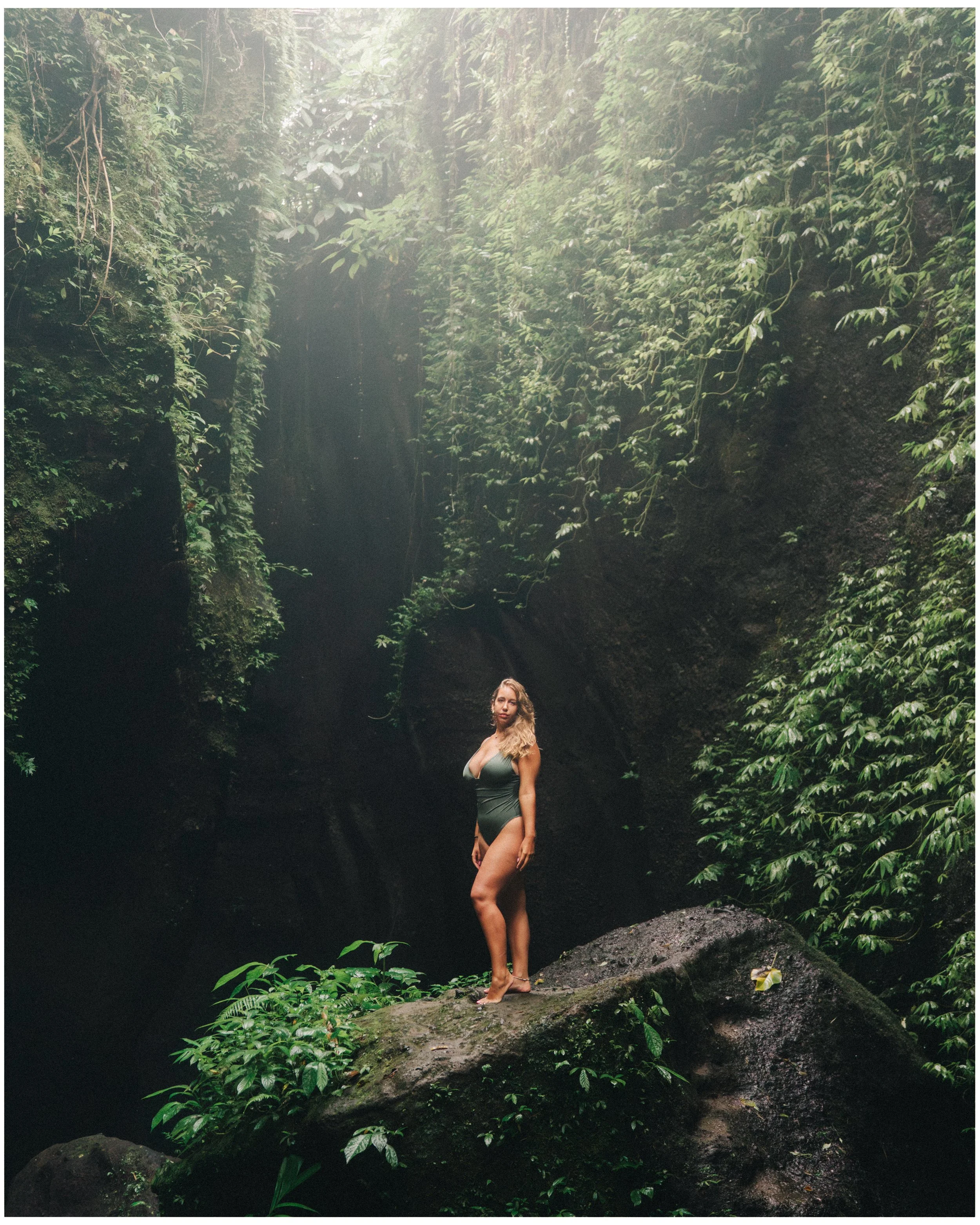 A woman in a green swimsuit standing barefoot on a rock in a lush, green jungle with dense foliage and sunlight filtering through the trees.