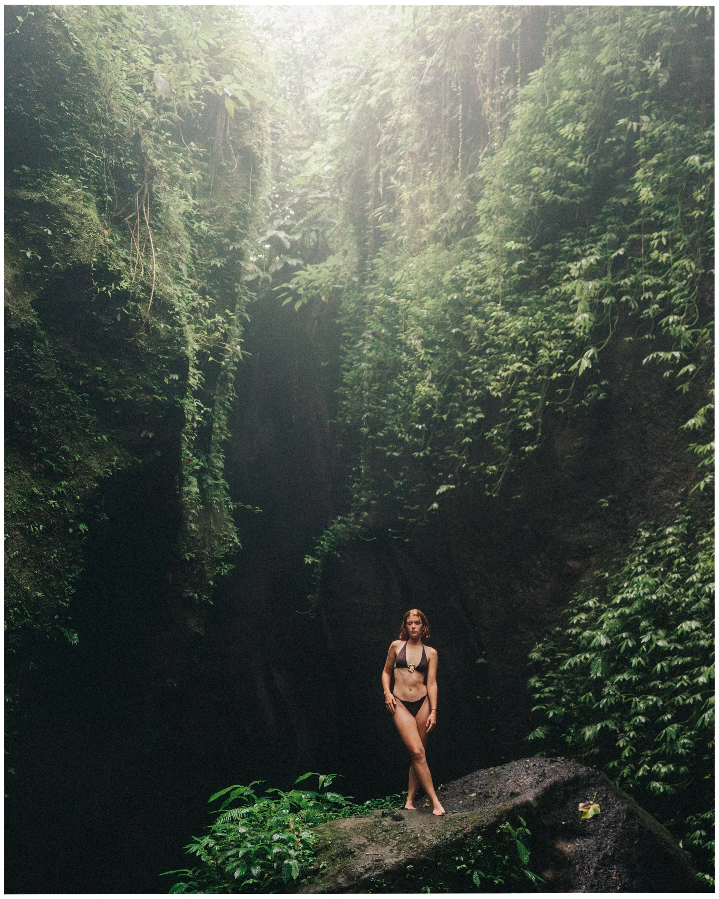 A woman in a black bikini standing on a large rock in a lush, green jungle or forest with sunlight coming through the trees.