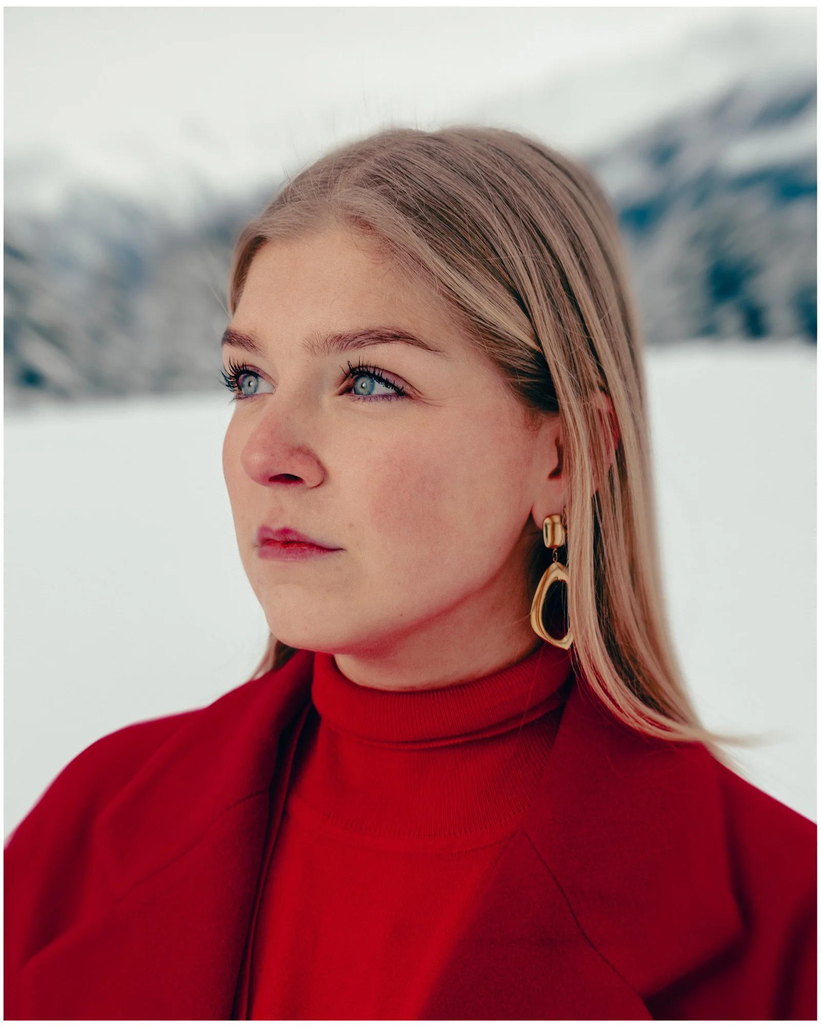 A young woman with blonde hair and blue eyes wearing a red turtleneck and gold earrings, standing outdoors in a snowy landscape with blurred trees in the background.