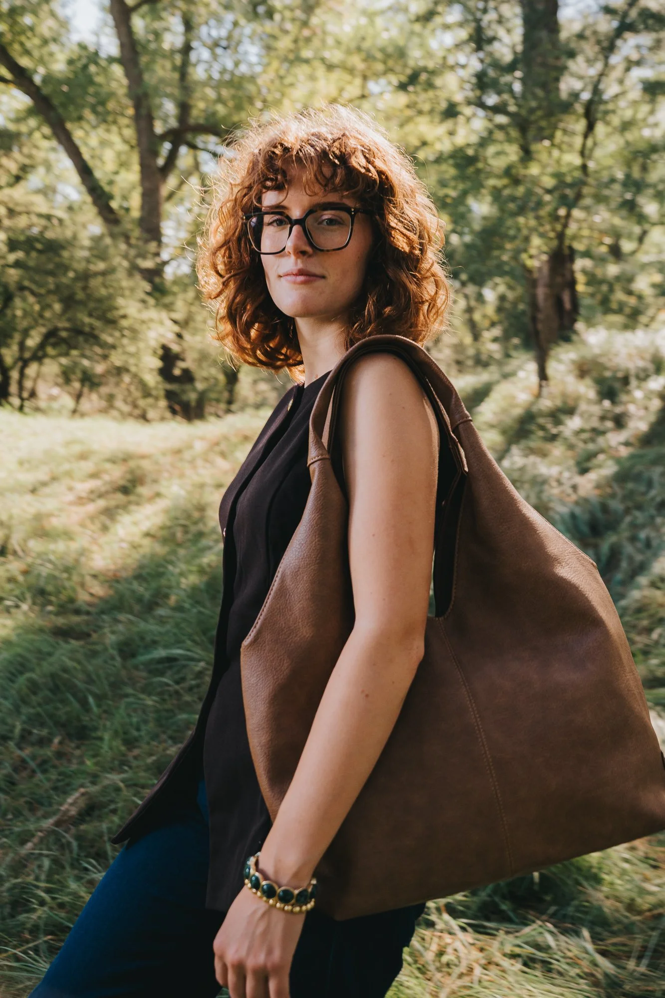 A young woman with curly red hair, glasses, and wearing a black sleeveless top, standing outdoors in a park-like setting with trees and grass, holding a large brown leather tote bag over her shoulder.