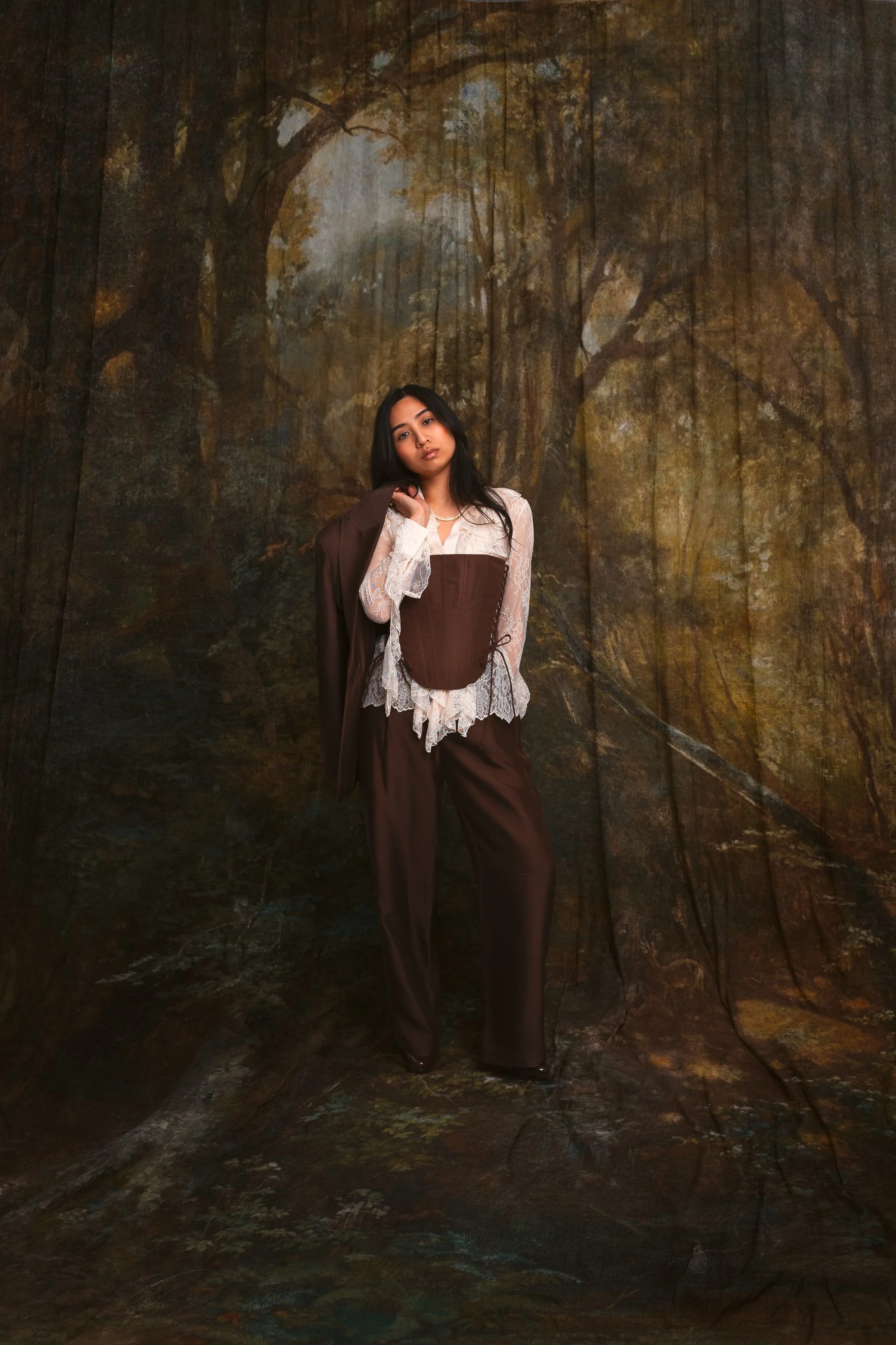 A woman standing indoors against a backdrop depicting a wooded forest scene. She wears a white lace blouse with a brown corset and matching brown pants, holding a brown jacket over her shoulder.
