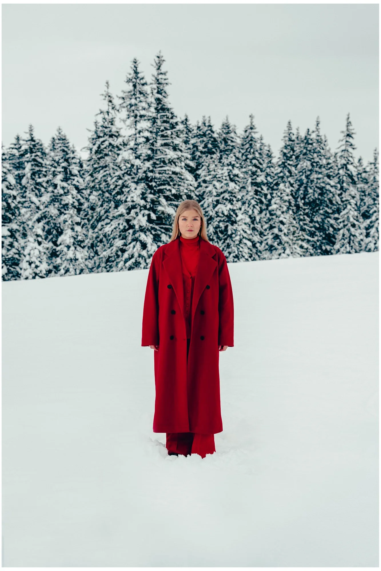 A woman in a red coat and pants standing in snow with a forest of snow-covered pine trees in the background.