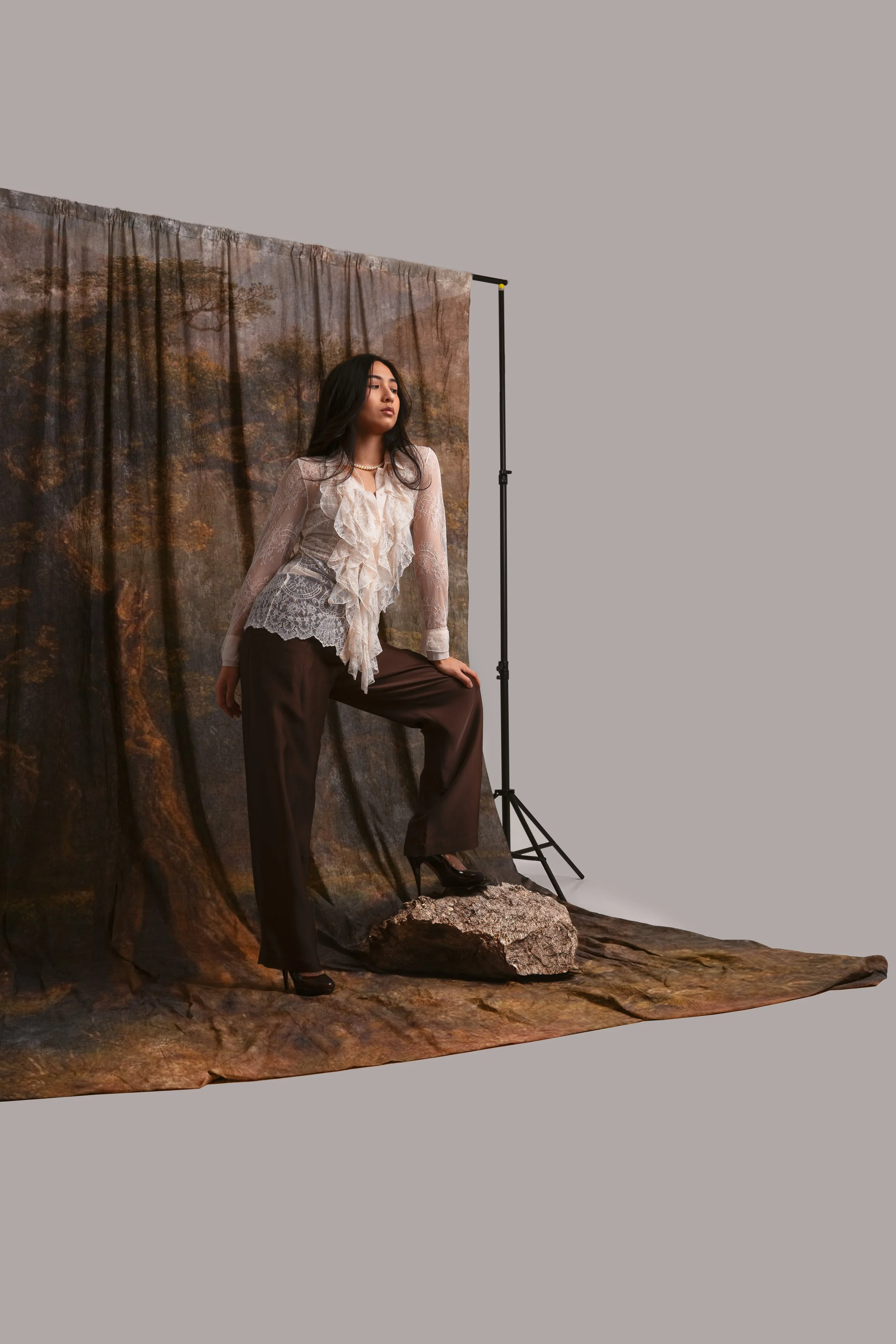 A woman in a vintage lace blouse and brown pants stands on a rocky surface in front of a autumn forest backdrop, with photography equipment visible.