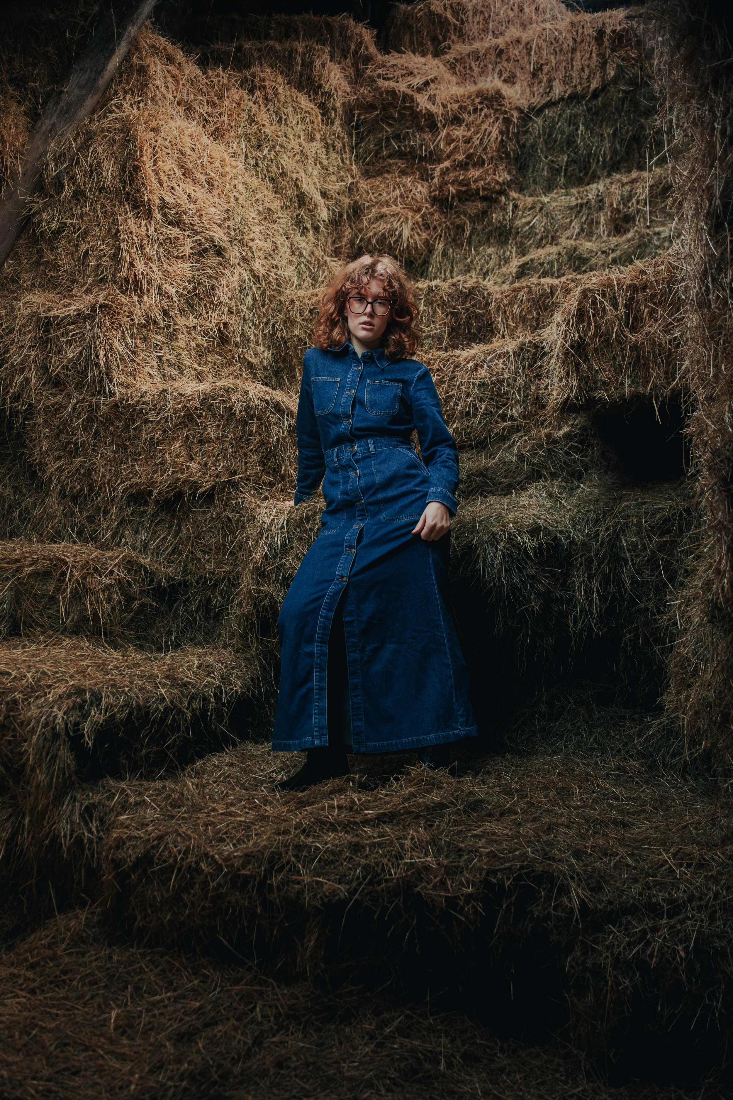 A woman wearing a blue denim dress and glasses poses in front of a background of stacked hay bales.