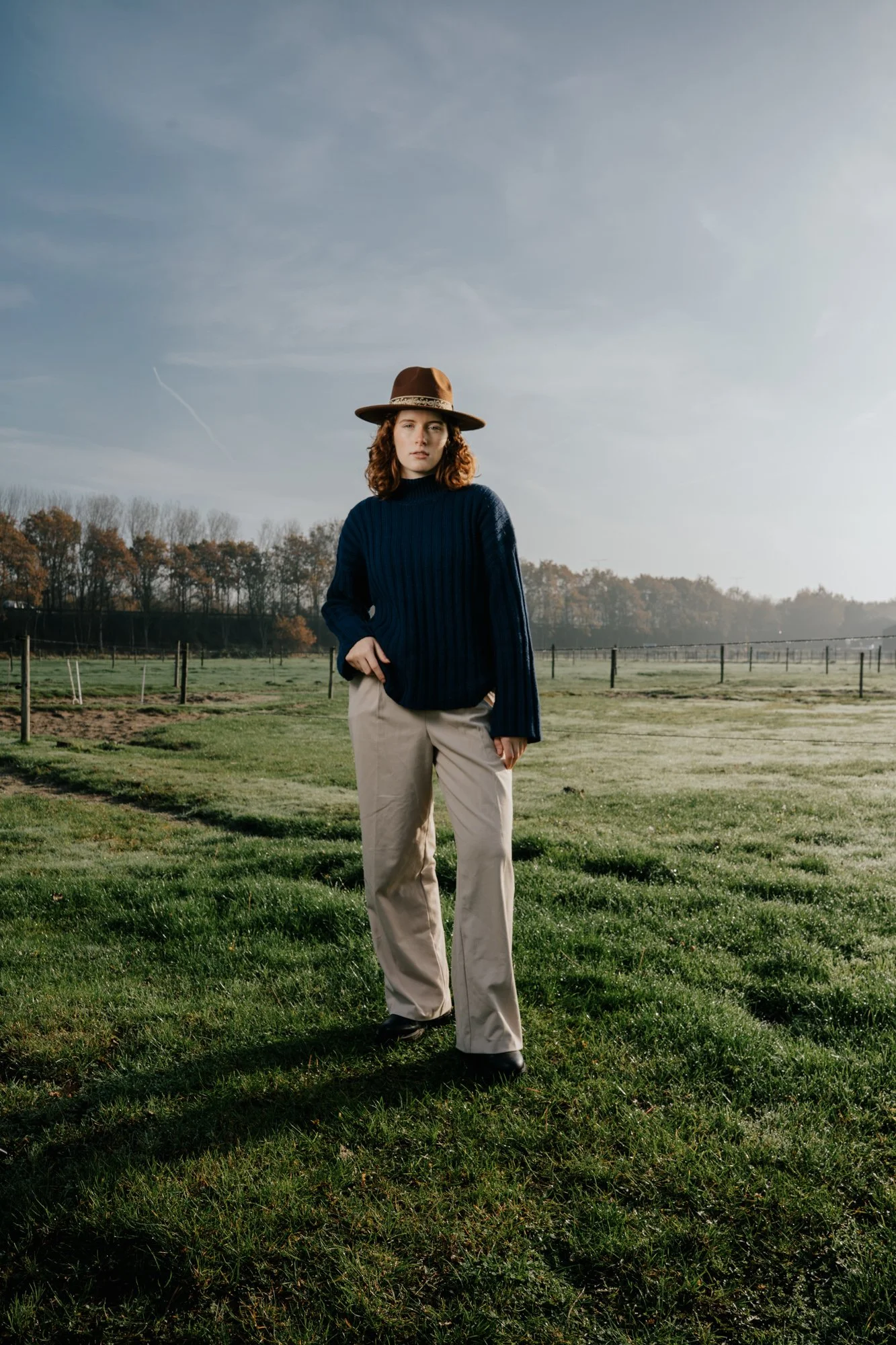A woman standing in a grassy field wearing a brown hat, navy blue sweater, and beige pants, with a cloudy sky and trees in the background.