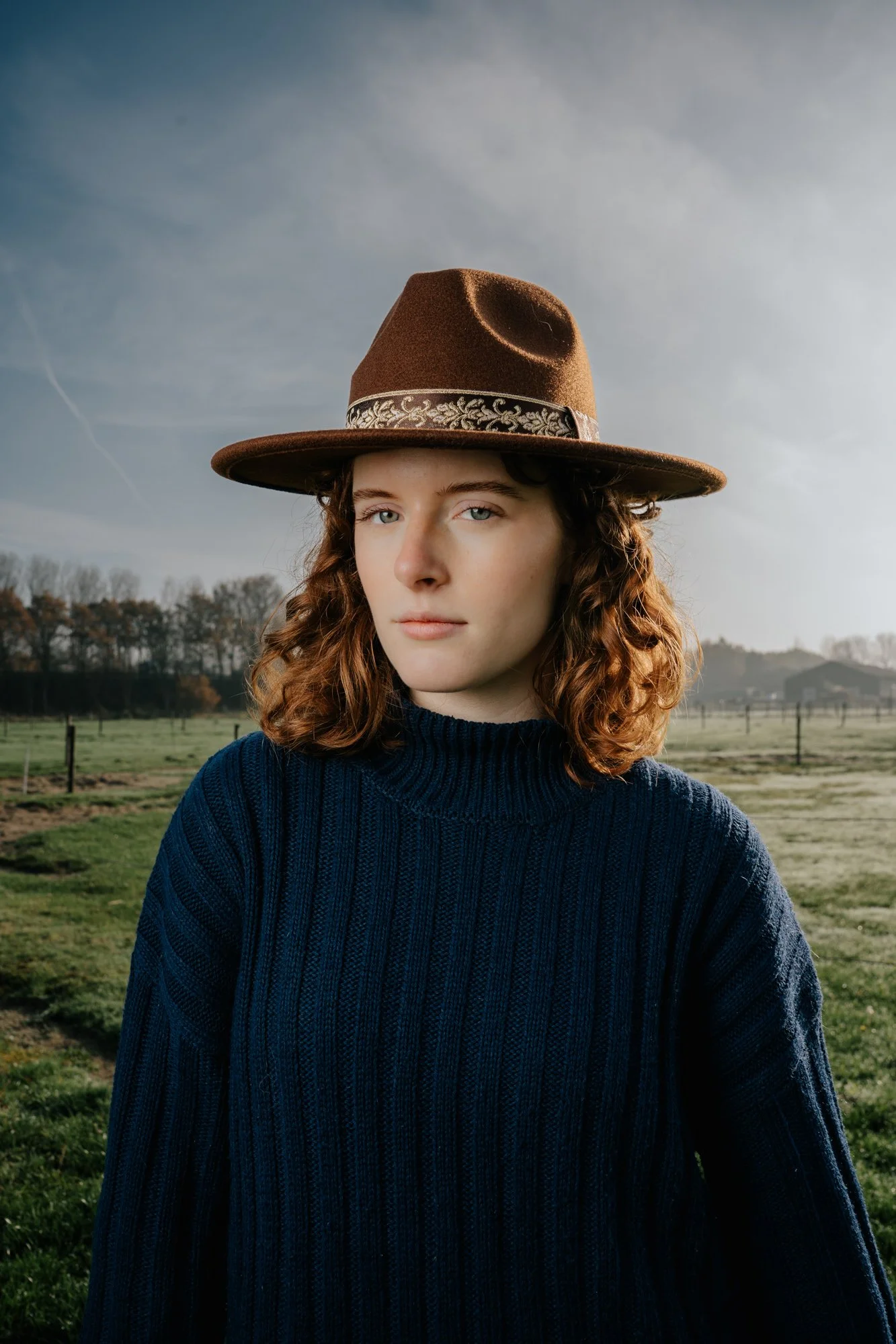 A woman with curly red hair wearing a navy blue knit sweater and a brown wide-brimmed hat standing outdoors in a grassy field with trees and a cloudy sky in the background.