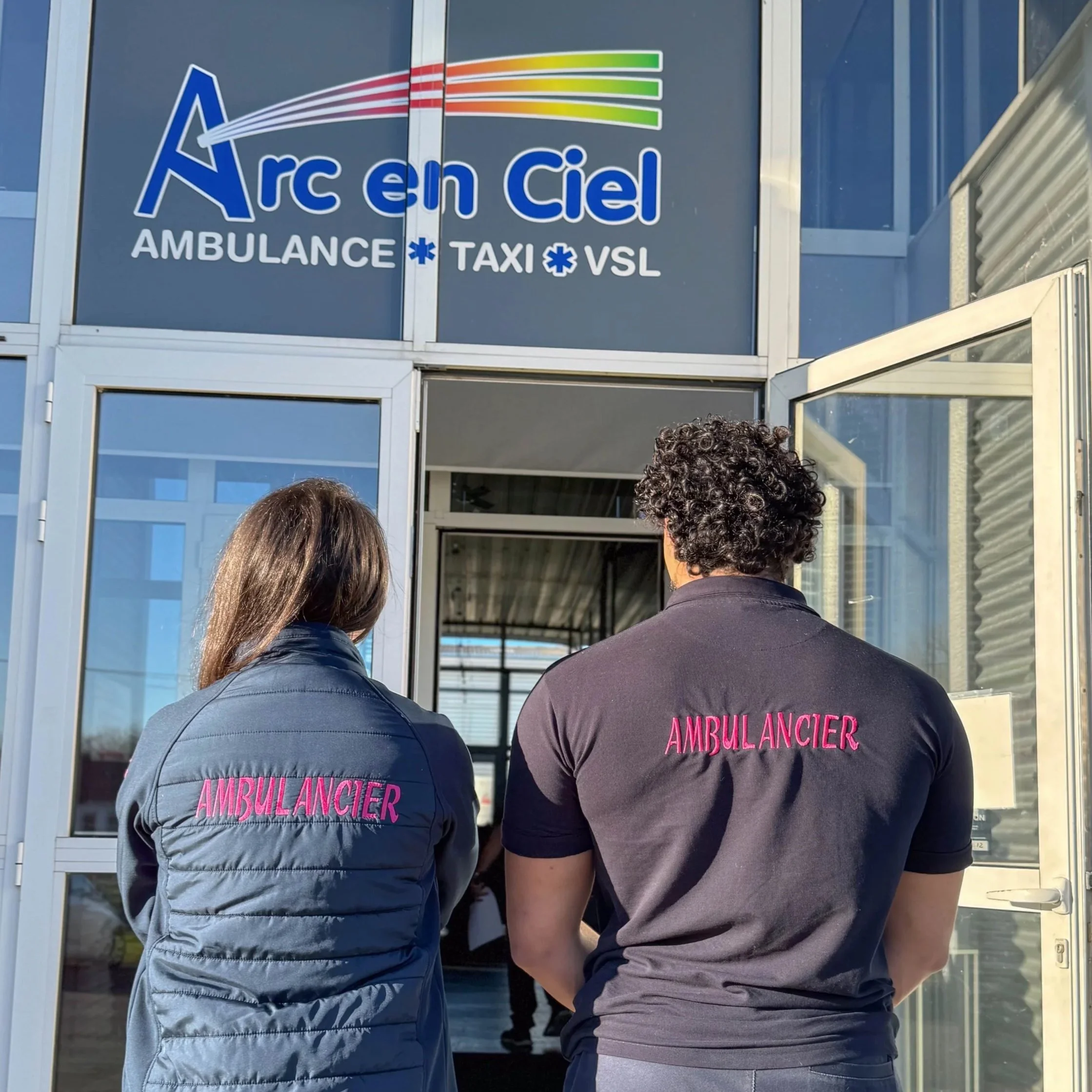 Deux personnes, dont une porte un veston avec 'AMBULANCIER' écrit en rose sur le dos, attendent devant une porte en verre, probablement à une ambulance ou une station de secours.