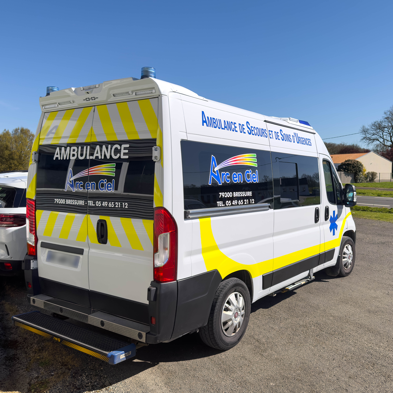 Véhicule d'urgence ambulance blanc avec des rayures jaunes et bleues, stationné en extérieur sous un ciel bleu, avec des bâtiments résidentiels en arrière-plan.