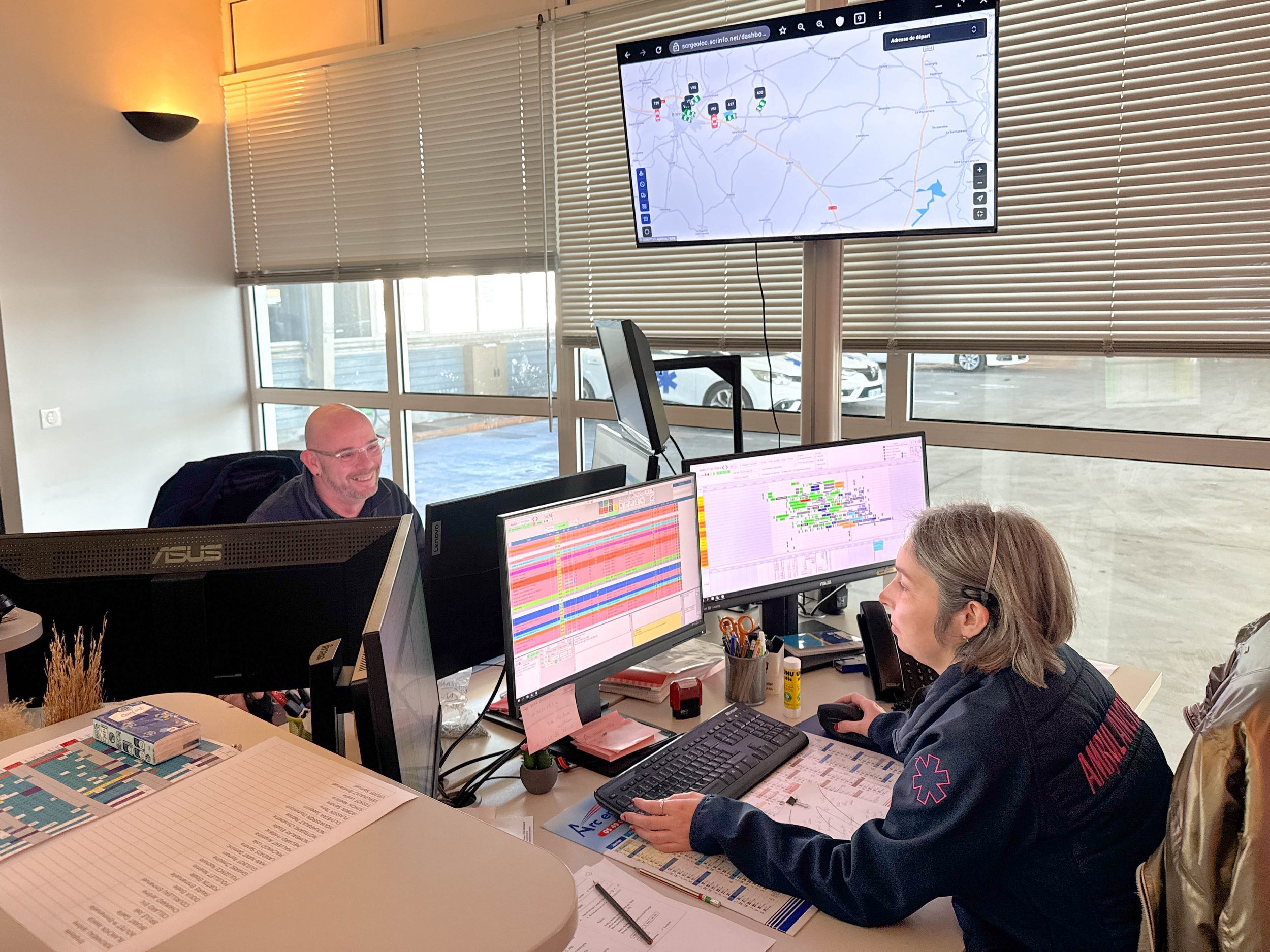 Un bureau avec plusieurs personnes travaillant devant des écrans d'ordinateur. Sur un grand écran suspendu au mur, une carte de localisation avec plusieurs icônes et une trajectoire est visible. La pièce est lumineuse avec des fenêtres en arrière-plan et des stores vénitiens.