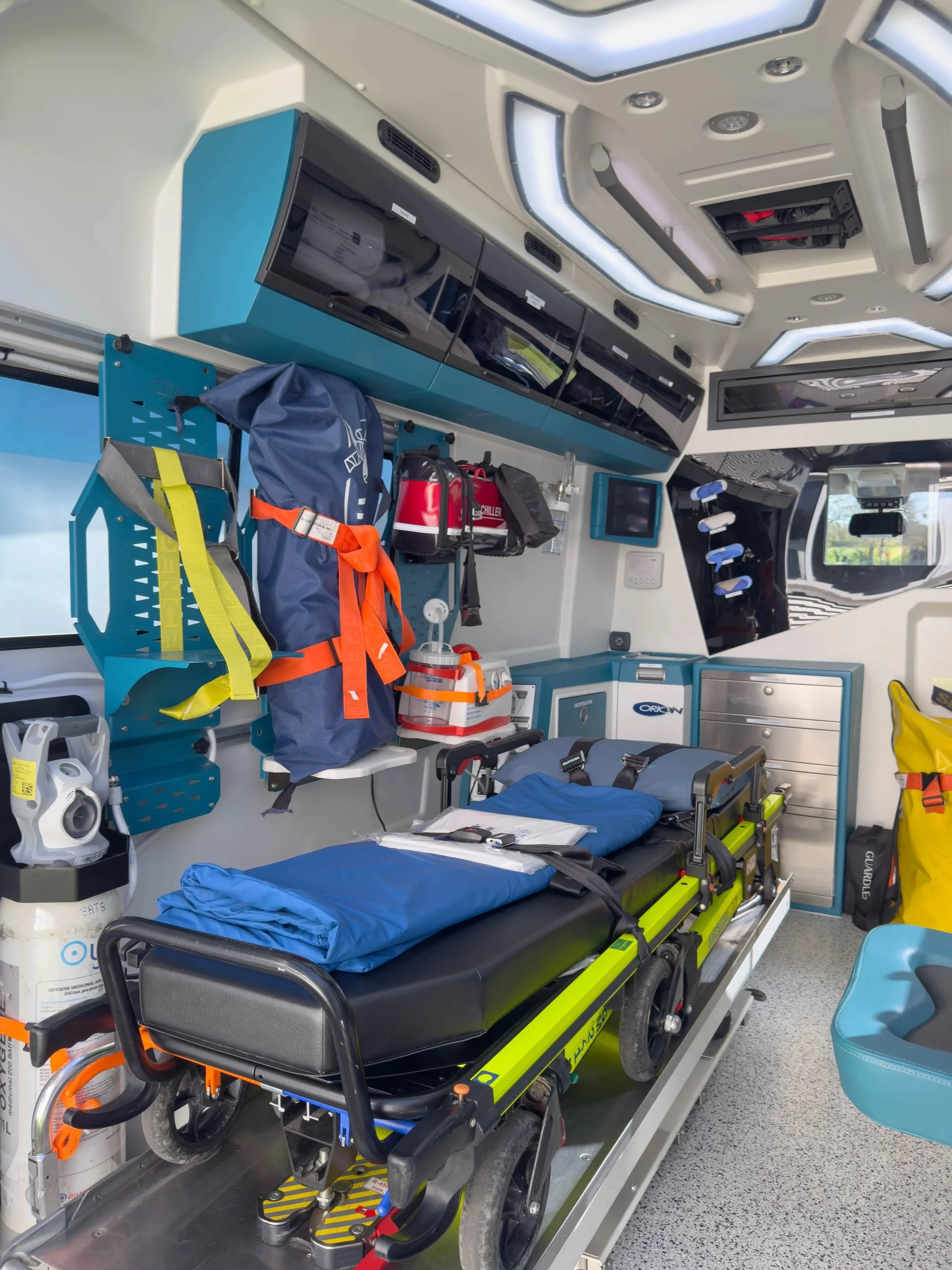 Intérieur d'une ambulance équipée avec un lit d'hôpital, des équipements médicaux, des sacs de secours, et appareils de surveillance.