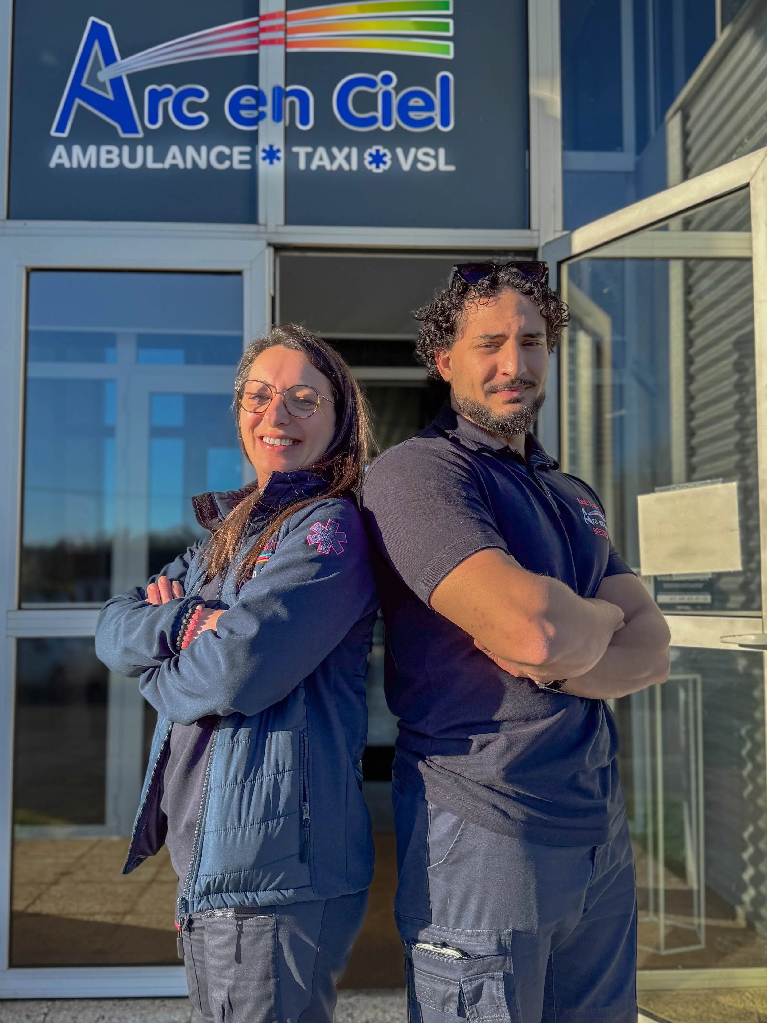 Deux ambulanciers, un homme et une femme, se tiennent dos à dos devant un bâtiment avec un panneau indiquant 'Arc en Ciel' et des services tels qu'ambulance, taxi et VSL, ensoleillé et soir.