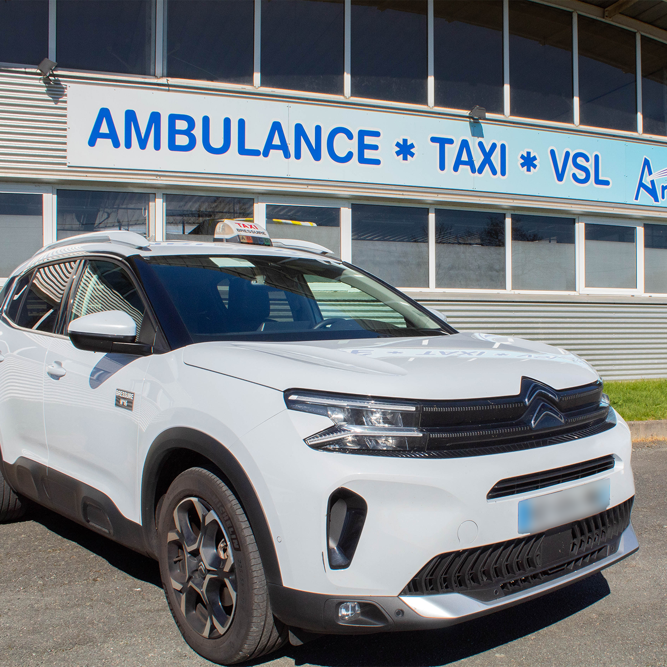 Voiture blanche de service avec un panneau indiquant ambuance, taxi et VSL devant un bâtiment avec de grandes fenêtres.
