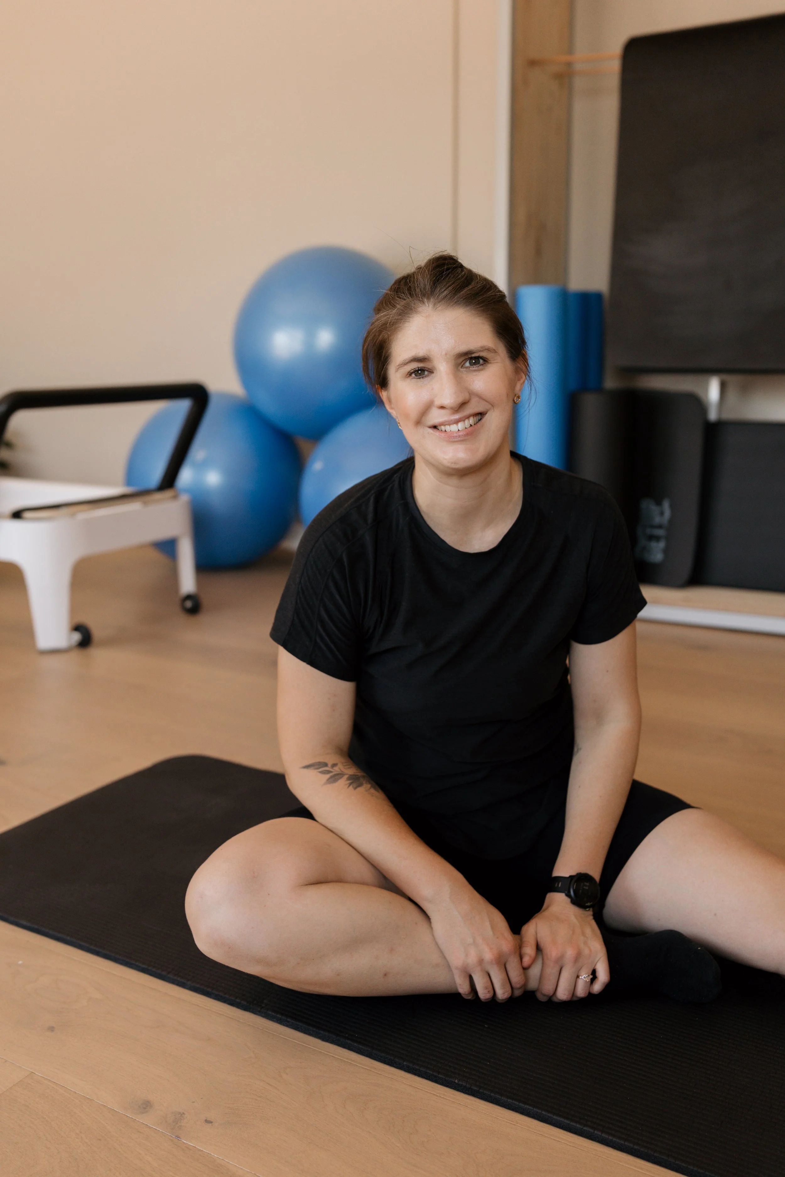 A woman sitting on a black yoga mat in a fitness studio, with blue exercise balls and foam rollers in the background.