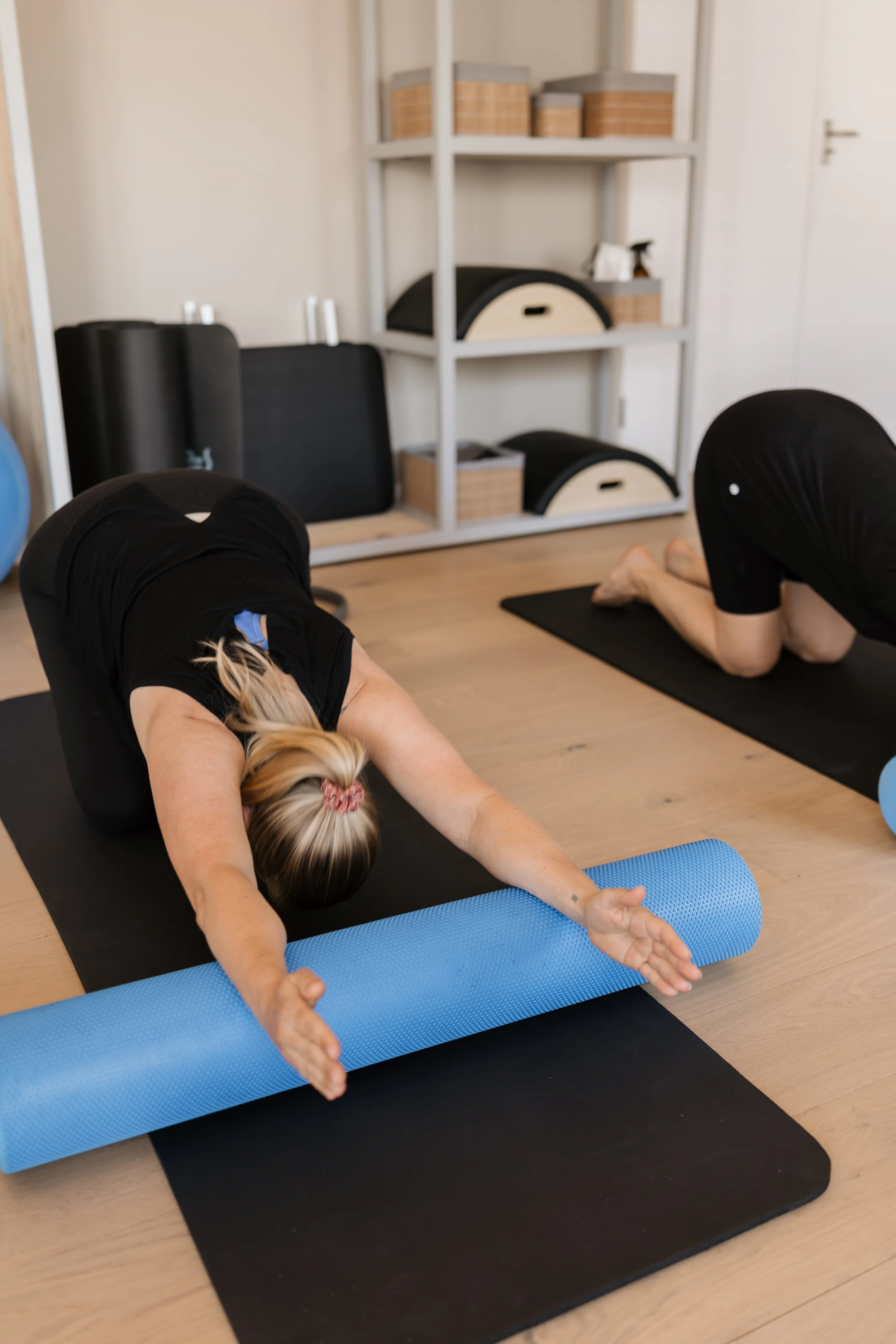 Two women practicing yoga on black mats, one with arms extended forward and the other with arms reaching overhead, in a room with shelving units and wooden flooring.