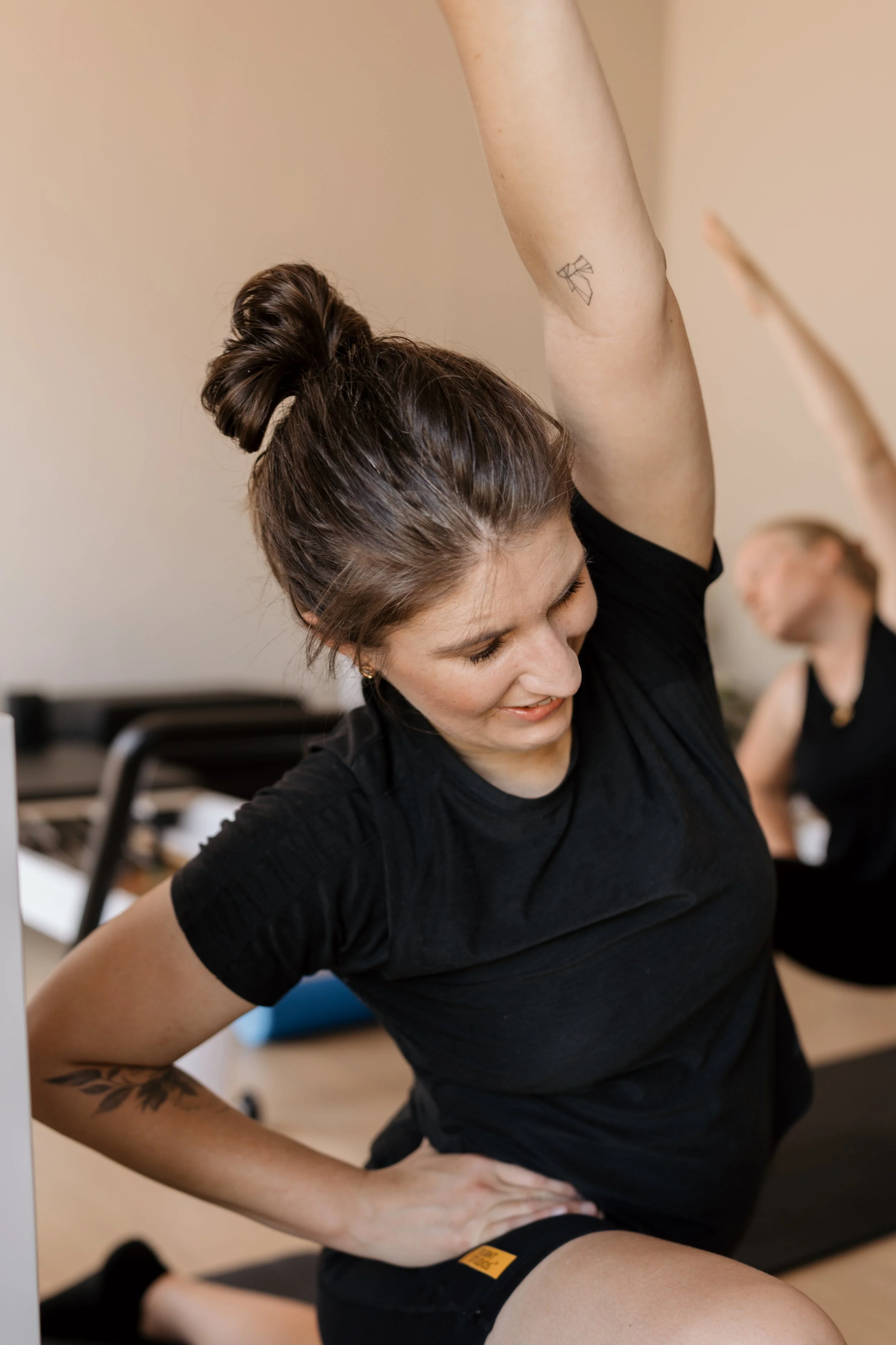 A woman with brown hair tied in a bun doing a stretching yoga pose in a room, wearing a black t-shirt and shorts, with a tattoo on her arm. Another person is blurred in the background.