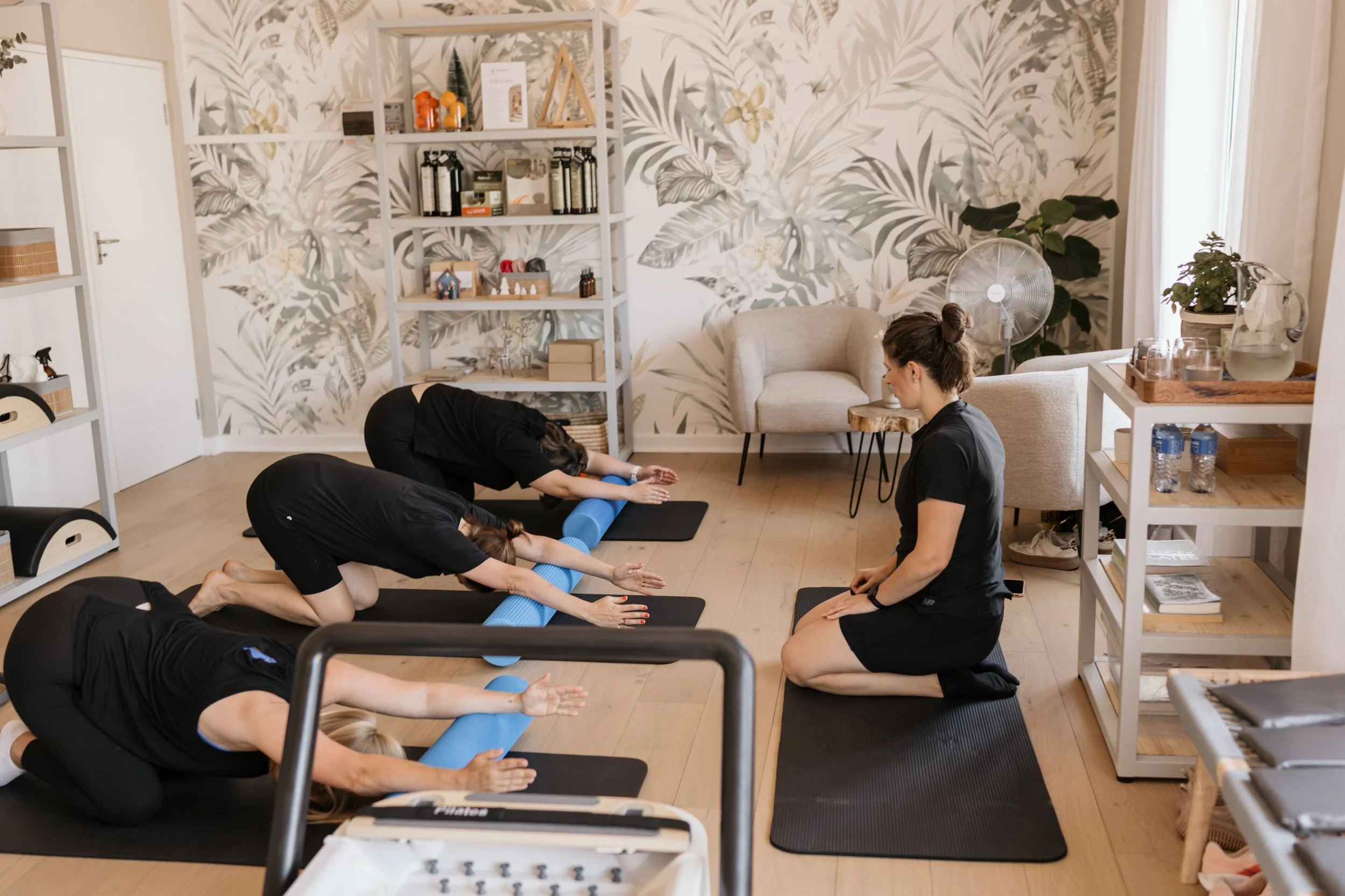People participating in a yoga class in a cozy room with botanical wallpaper, shelves with decorative items, and large windows.