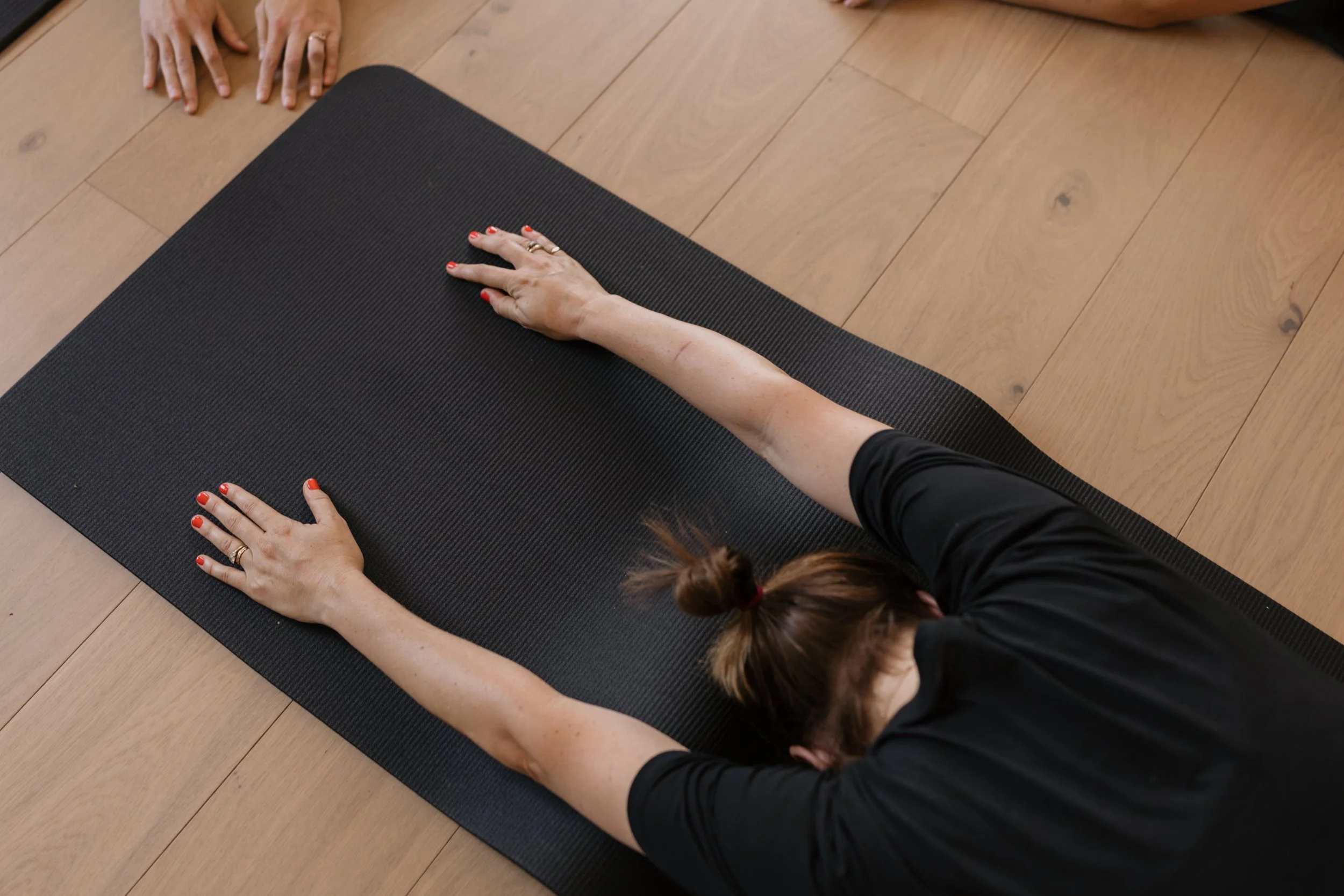 A person practicing yoga or stretching on a black mat, with their hands and forehead on the floor, wearing black clothing and red nail polish, on a wooden floor.