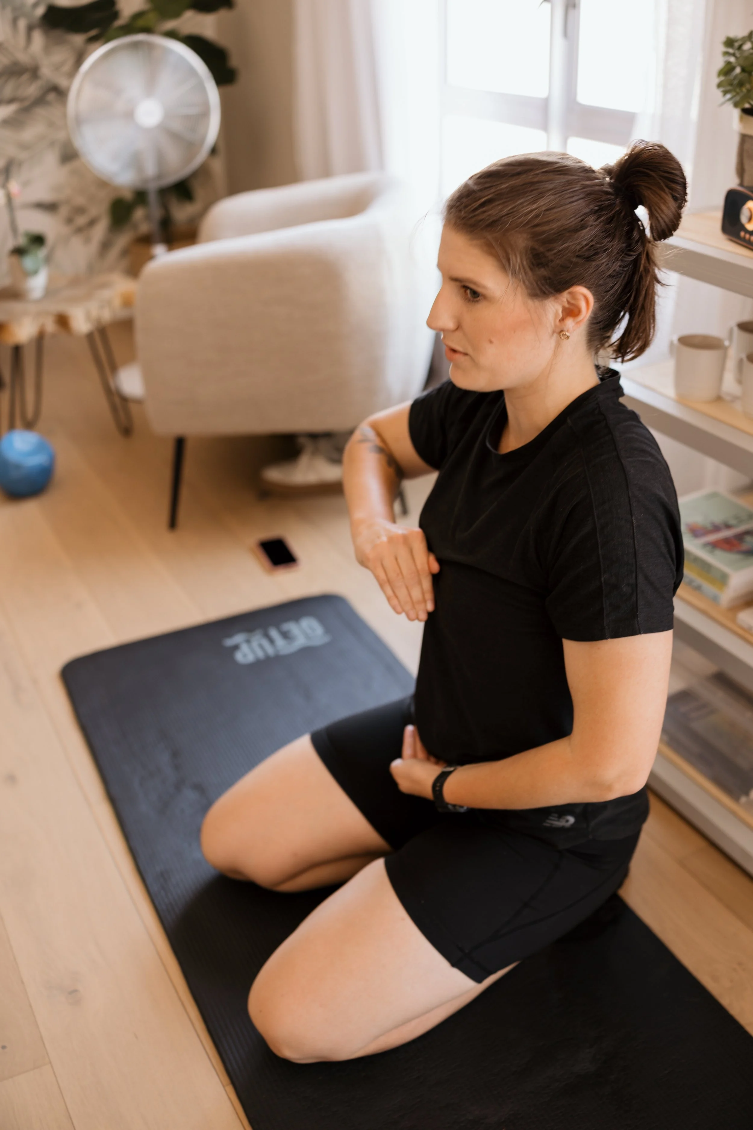 A woman with brown hair tied in a bun is kneeling on a black exercise mat, wearing a black t-shirt and black shorts, in a room with a wooden floor, a white shelf with mugs, a fan, a window with curtains, and a dining area in the background.