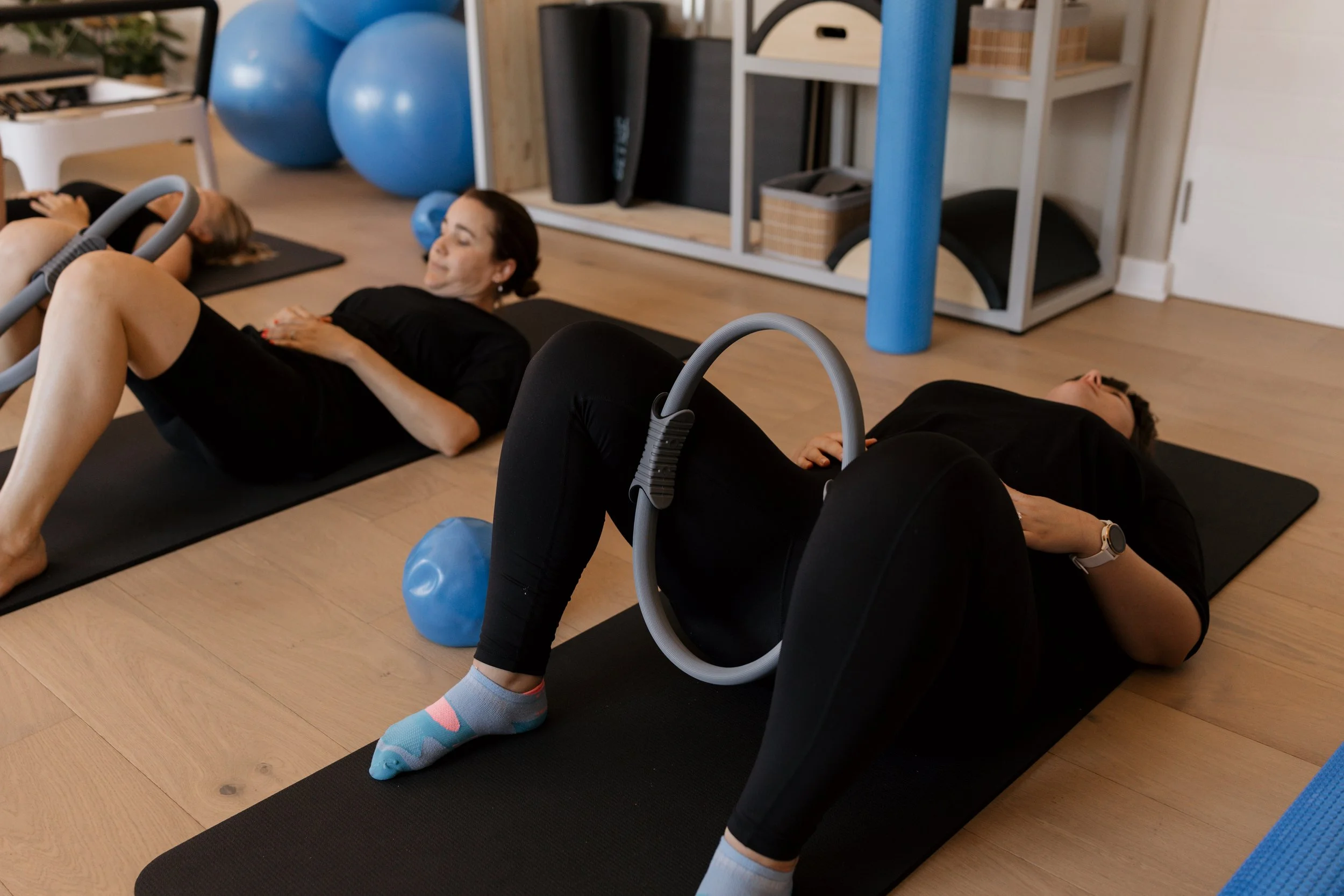 Two women lying on black exercise mats, engaging in core exercises with blue exercise balls and gray resistance bands in a well-equipped gym or fitness studio.