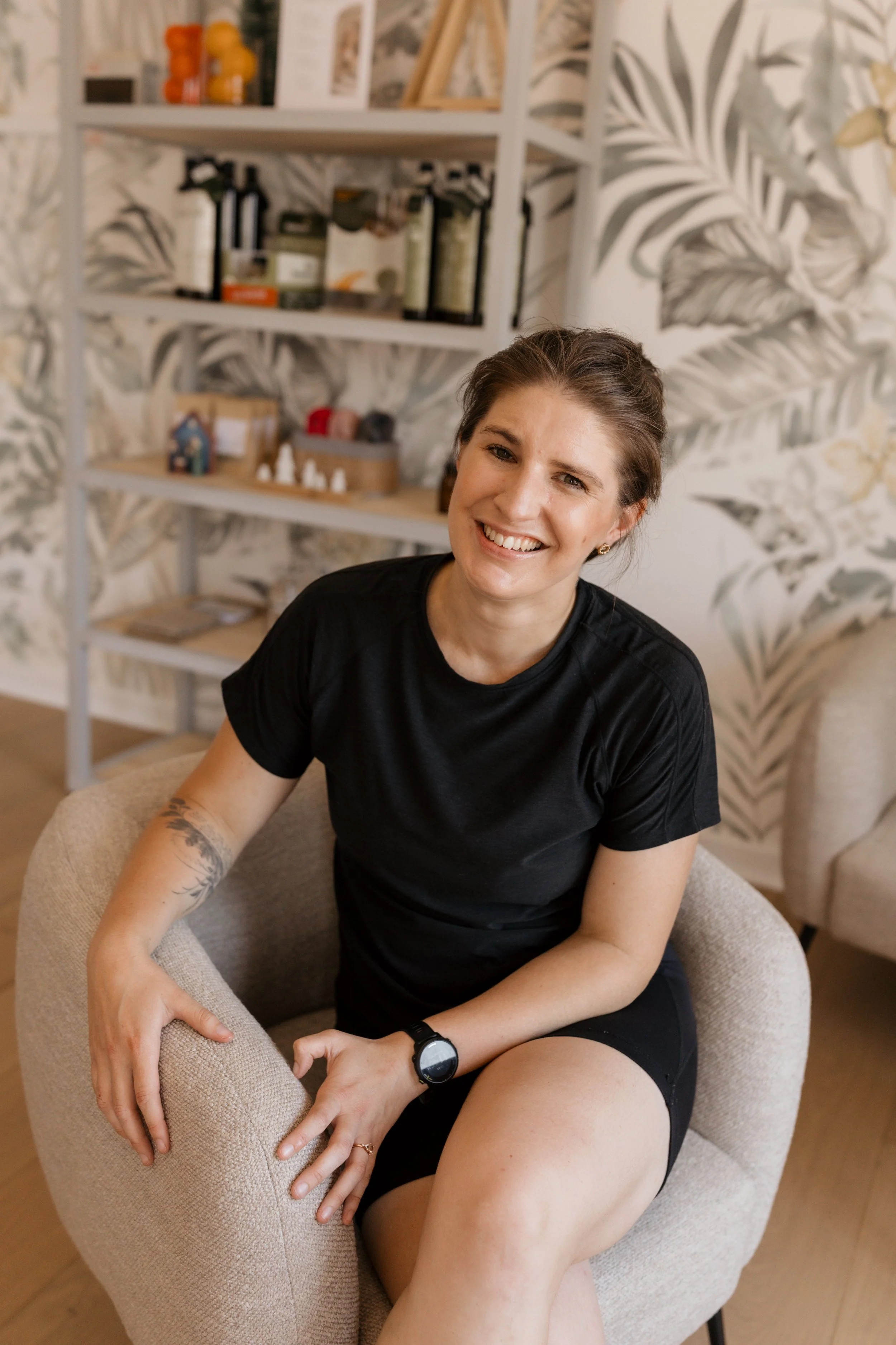 A woman with a big smile, wearing a black T-shirt and shorts, sitting comfortably in a beige armchair in a cozy room with a patterned wallpaper background. She has short brown hair, tattoos on her left arm, and a watch on her left wrist.