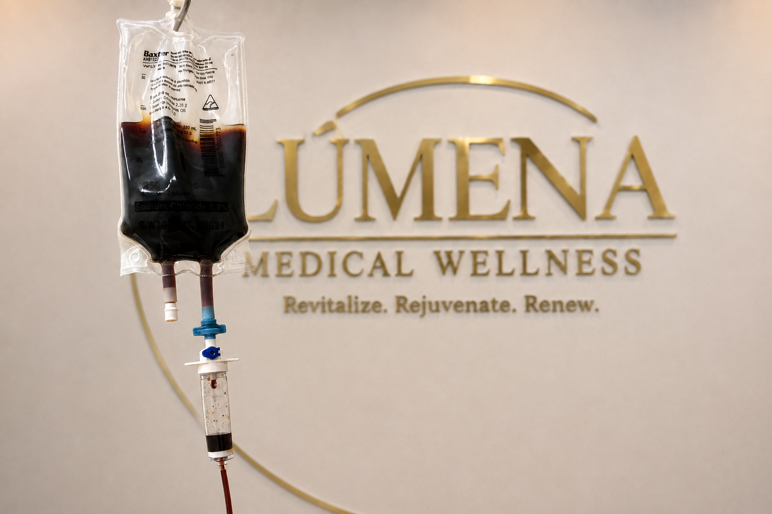 Intravenous infusion bag containing dark red liquid hanging in front of a wall with 'LUMENA Medical Wellness' logo and slogan 'Revitalize. Rejuvenate. Renew.'