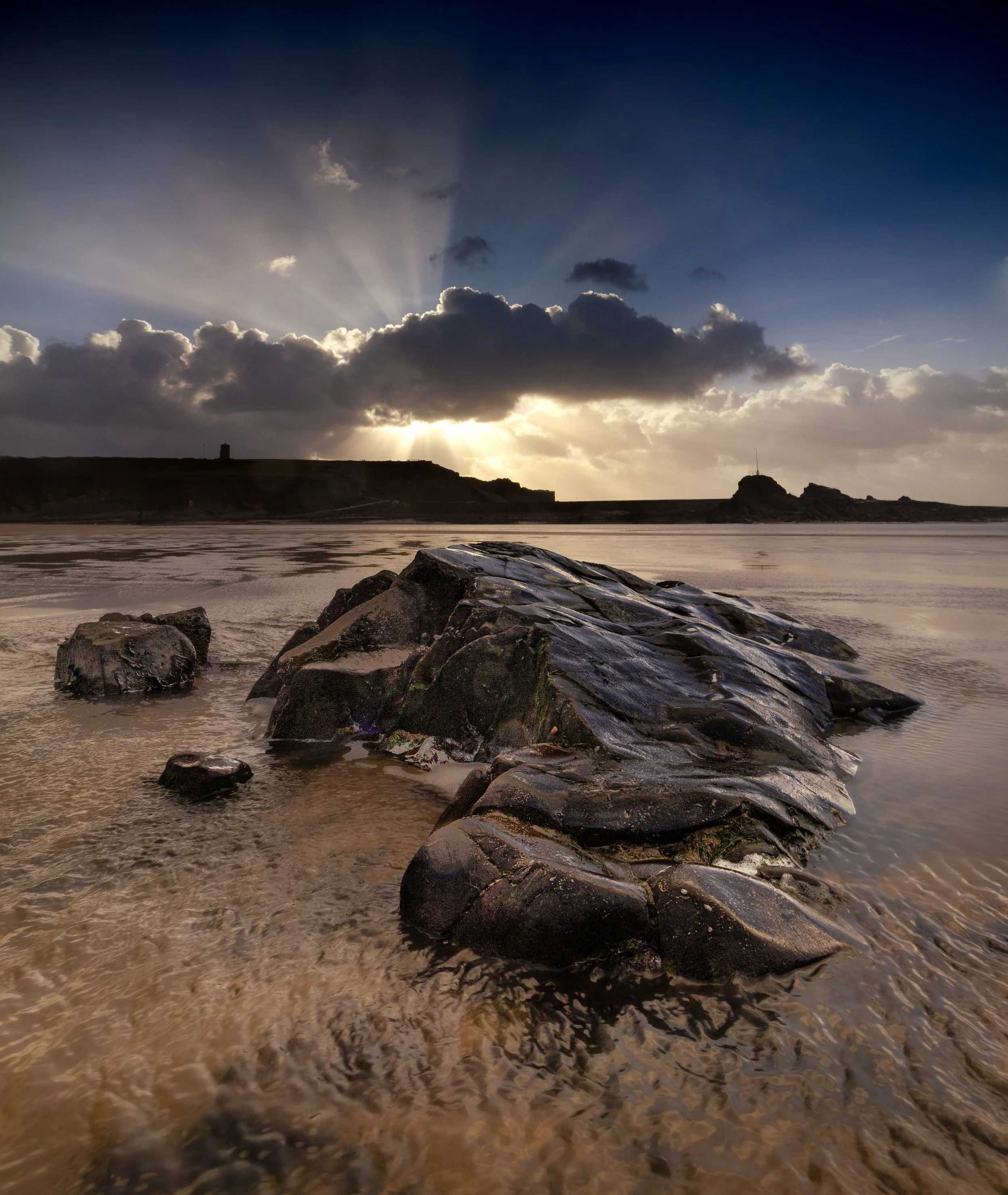 Sunset over a beach with large black rocks and calm water in foreground, with a cloudy sky and sun rays breaking through clouds in background.