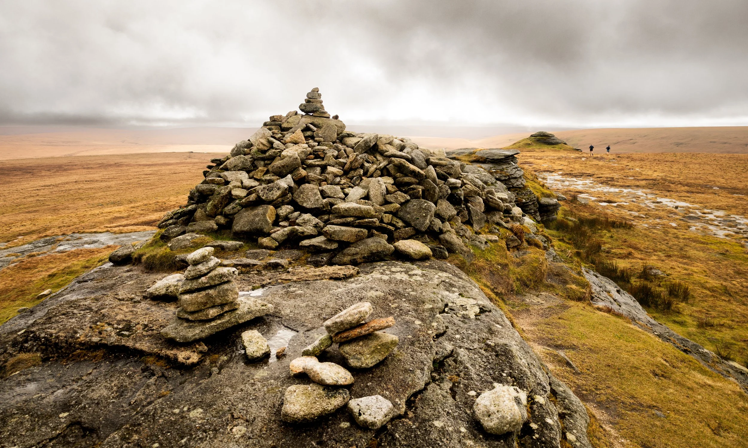 A landscape of open moorland with a large pile of stones in the foreground and two people walking in the distance under a cloudy sky.